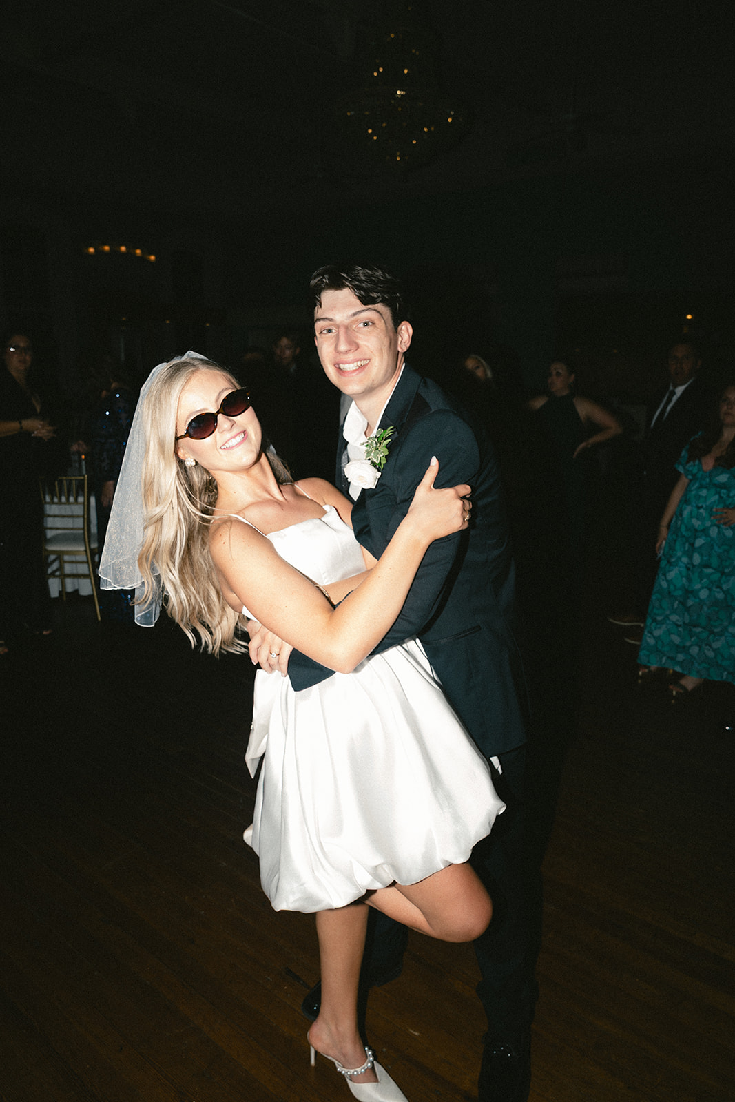 A bride in a short white dress and veil walks beside a groom in a black suit; both wear sunglasses and hold hands on a sidewalk at night.