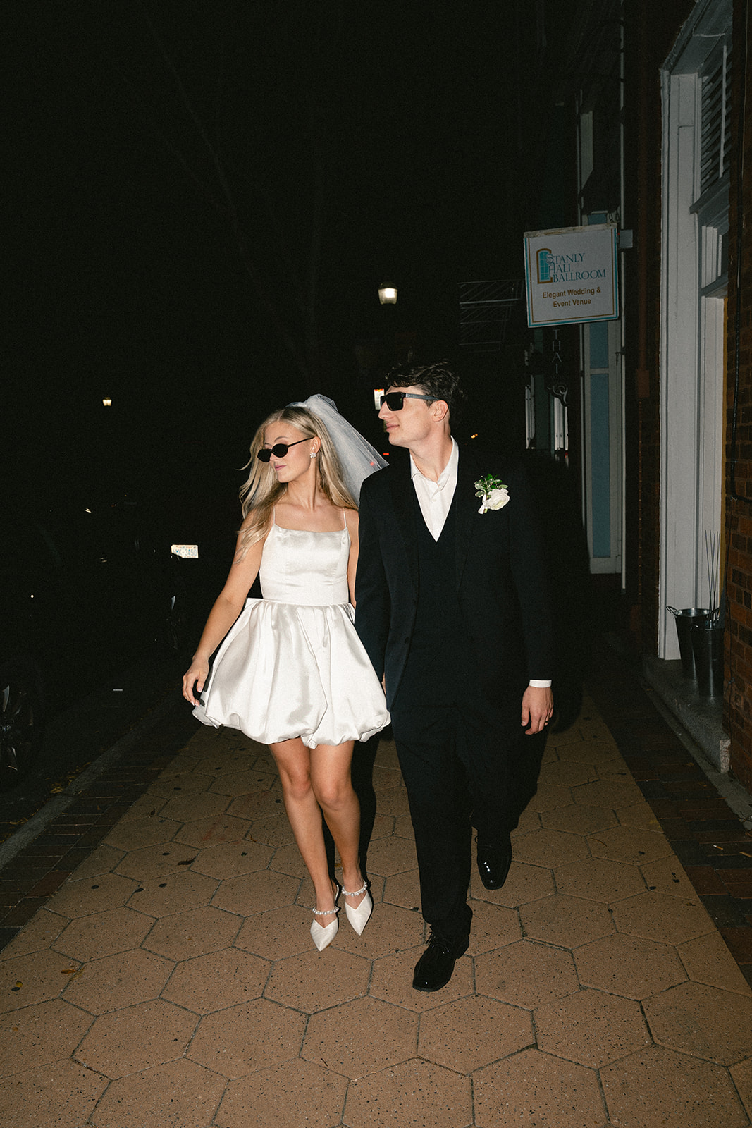 A bride in a short white dress and veil walks beside a groom in a black suit; both wear sunglasses and hold hands on a sidewalk at night.