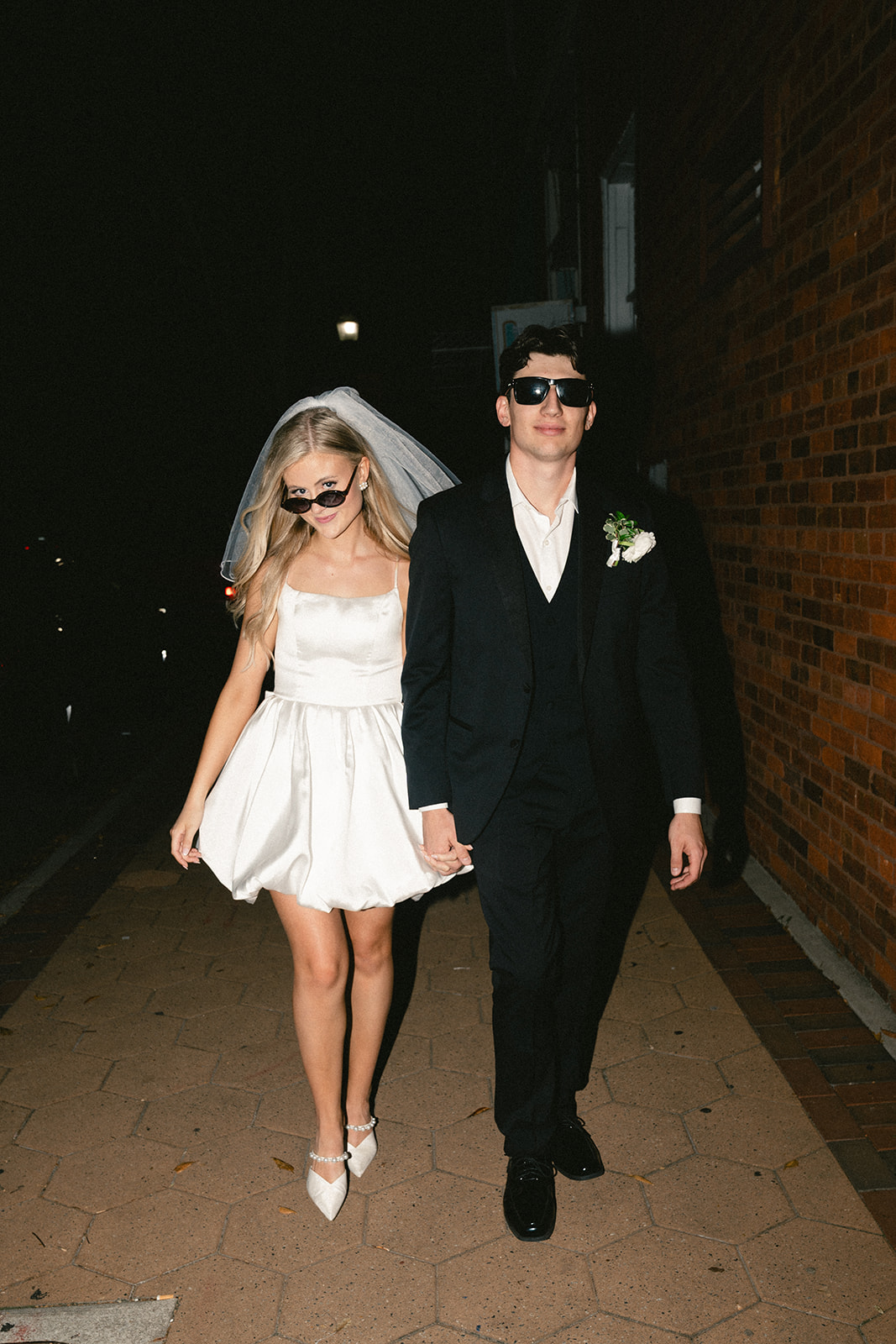 A bride in a short white dress and veil walks beside a groom in a black suit; both wear sunglasses and hold hands on a sidewalk at night.