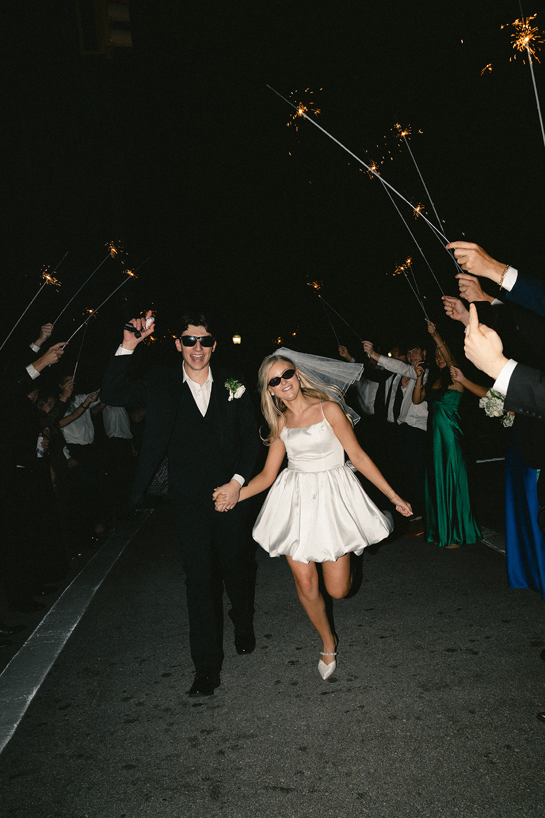 A bride in a short white dress and veil walks beside a groom in a black suit; both wear sunglasses and hold hands on a sidewalk at night.