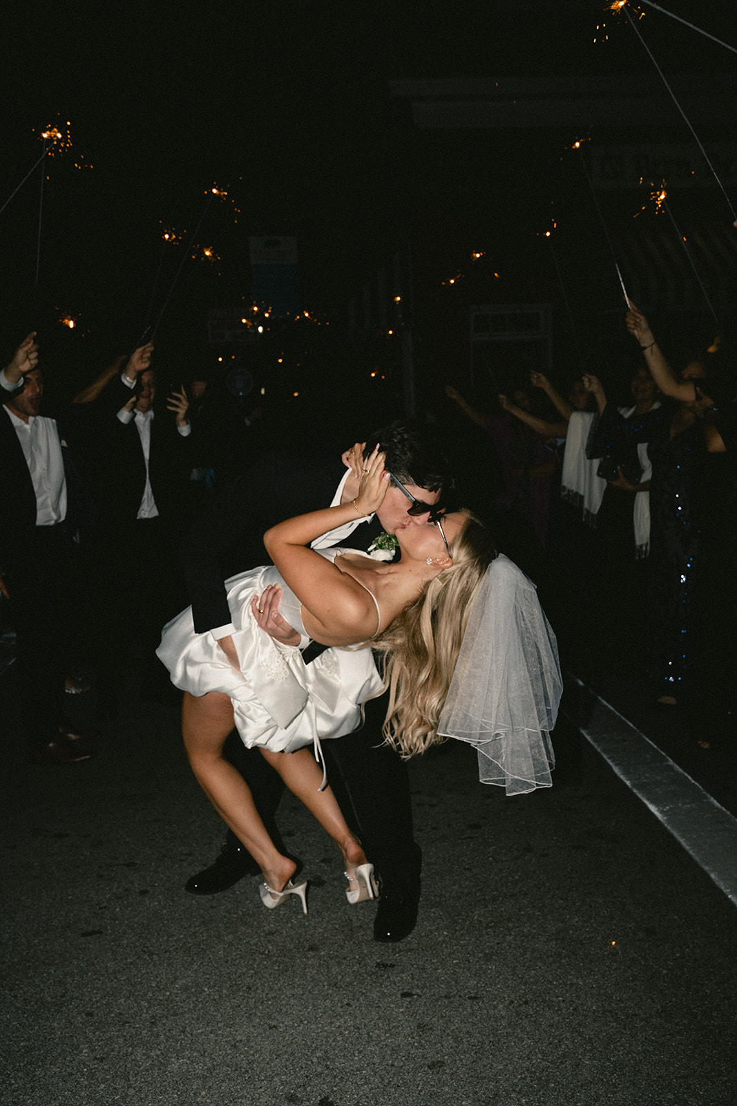 A bride in a short white dress and veil walks beside a groom in a black suit; both wear sunglasses and hold hands on a sidewalk at night.