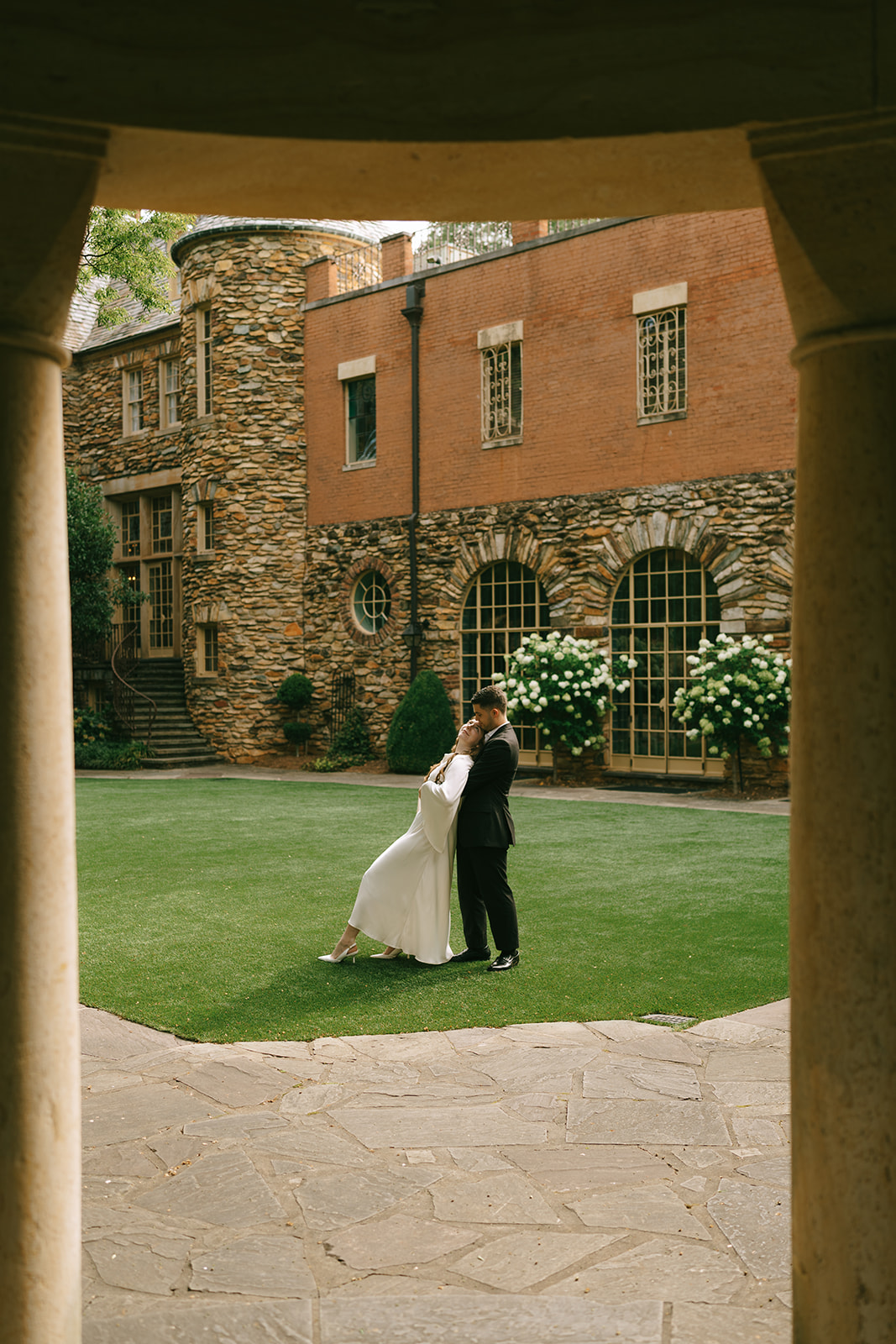 A woman in a white dress and a man in a black suit hold hands while walking on a cobblestone path in front of a stone building. | What to Wear for Engagement Photos