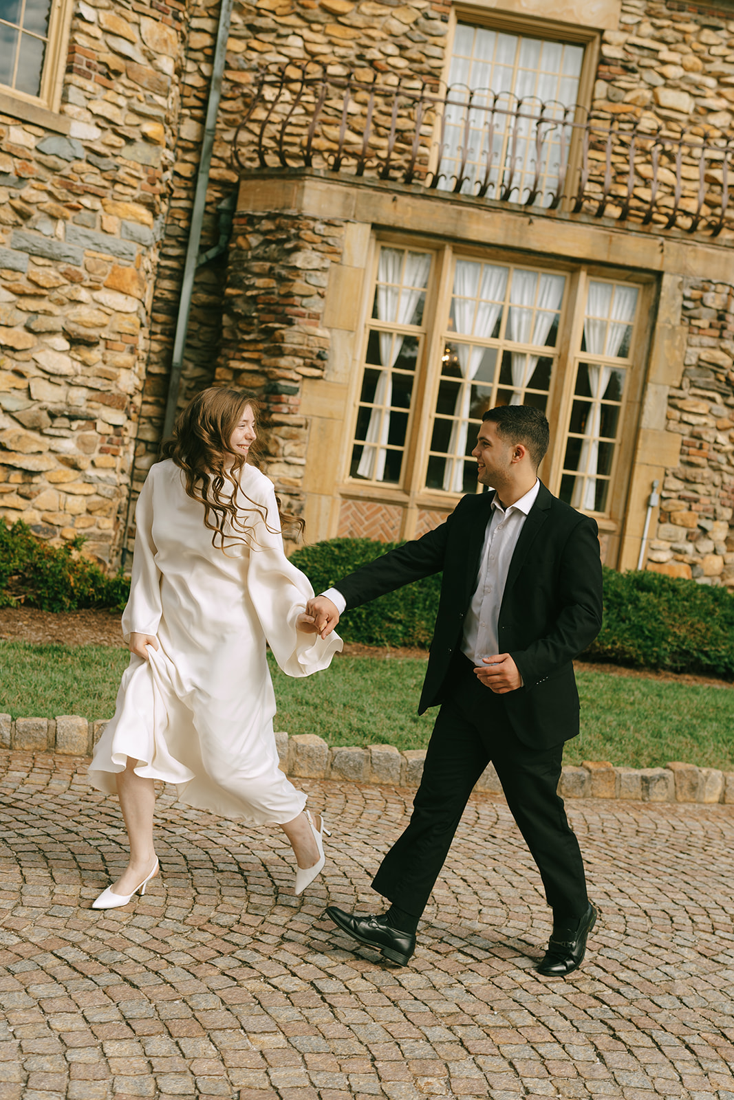 A woman in a white dress and a man in a black suit hold hands while walking on a cobblestone path in front of a stone building. | What to Wear for Engagement Photos
