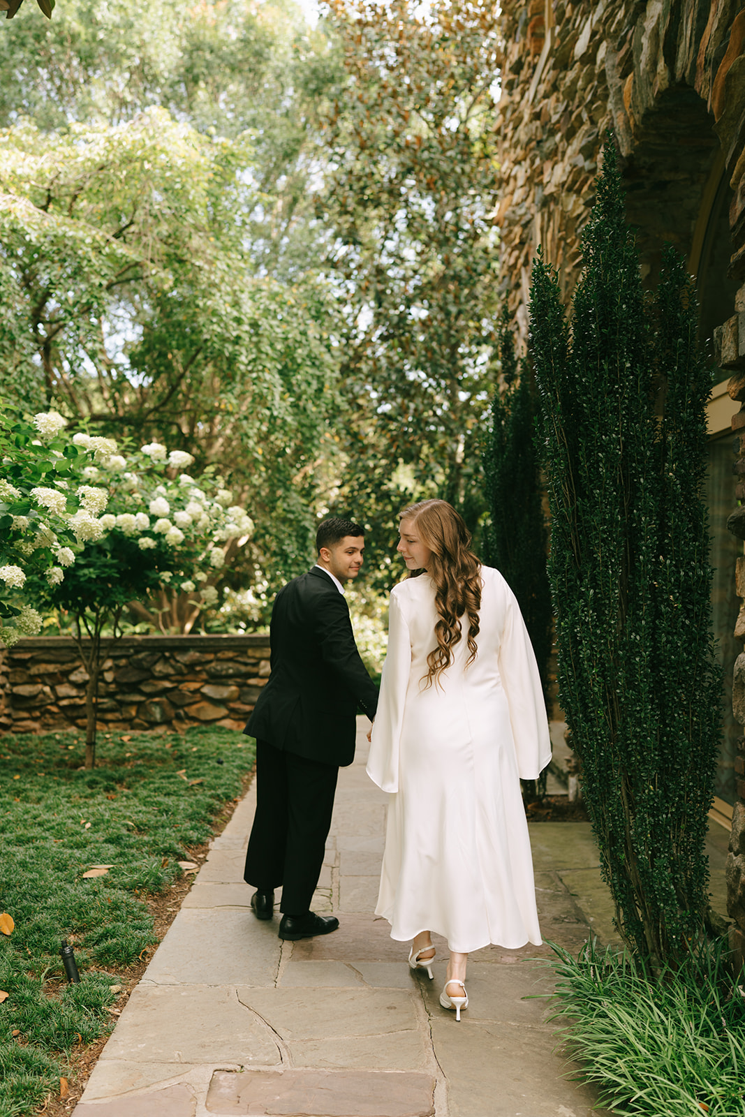 A woman in a white dress and a man in a black suit hold hands while walking on a cobblestone path in front of a stone building. | What to Wear for Engagement Photos
