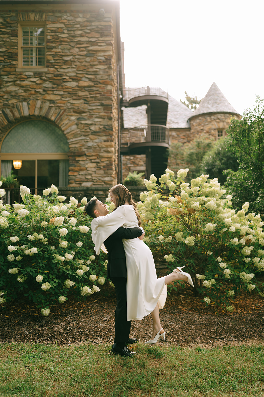 A newlywed couple poses in front of a stone building and blooming bushes; the groom lifts the bride, who smiles and holds up her leg.