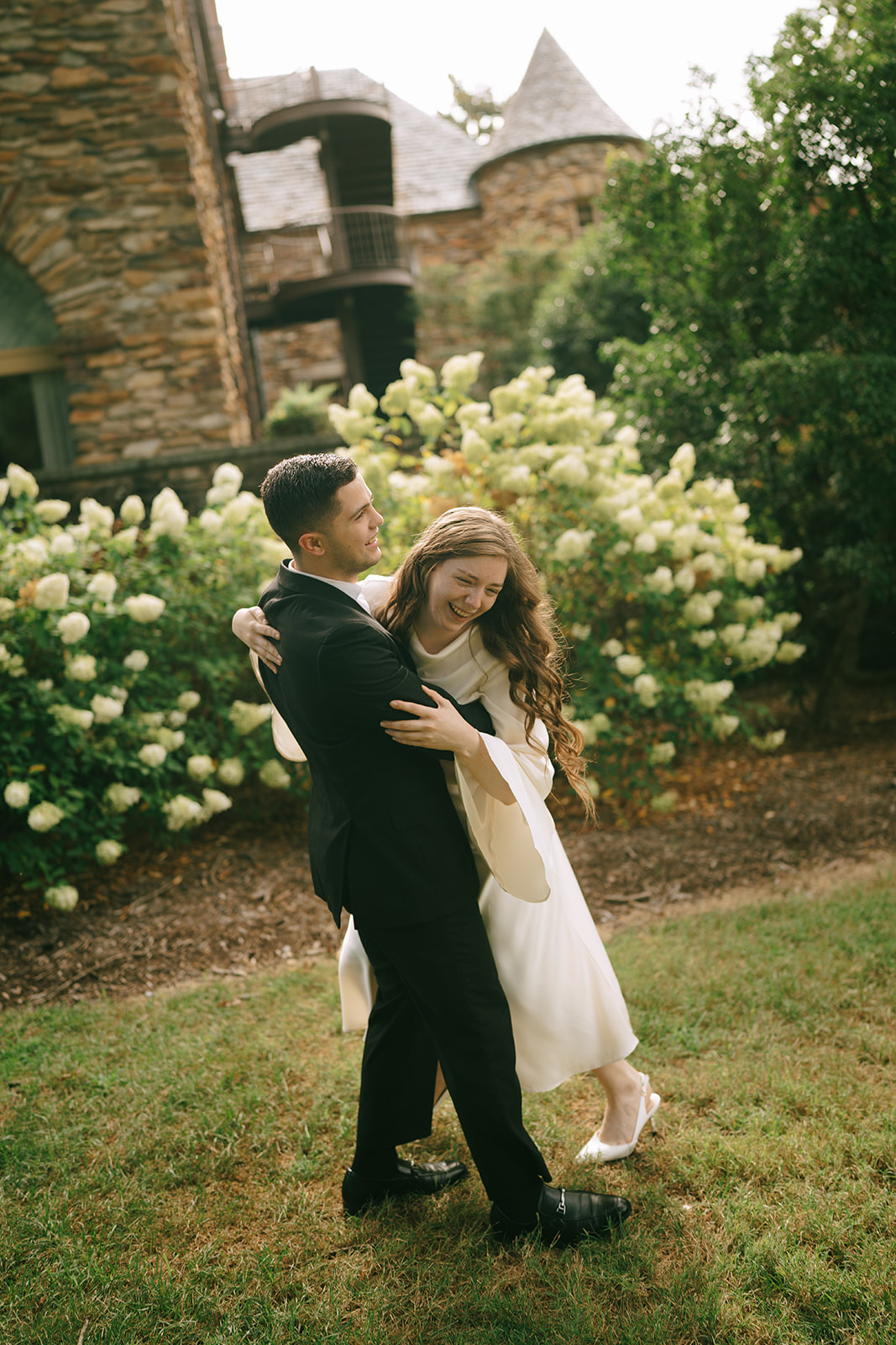 A newlywed couple poses in front of a stone building and blooming bushes; the groom lifts the bride, who smiles and holds up her leg.