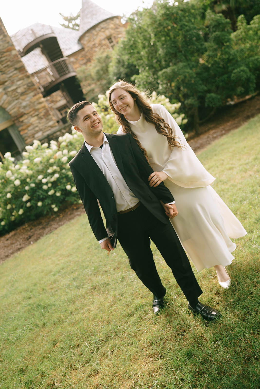 A woman in a white dress and a man in a black suit hold hands while walking on a cobblestone path in front of a stone building. | What to Wear for Engagement Photos