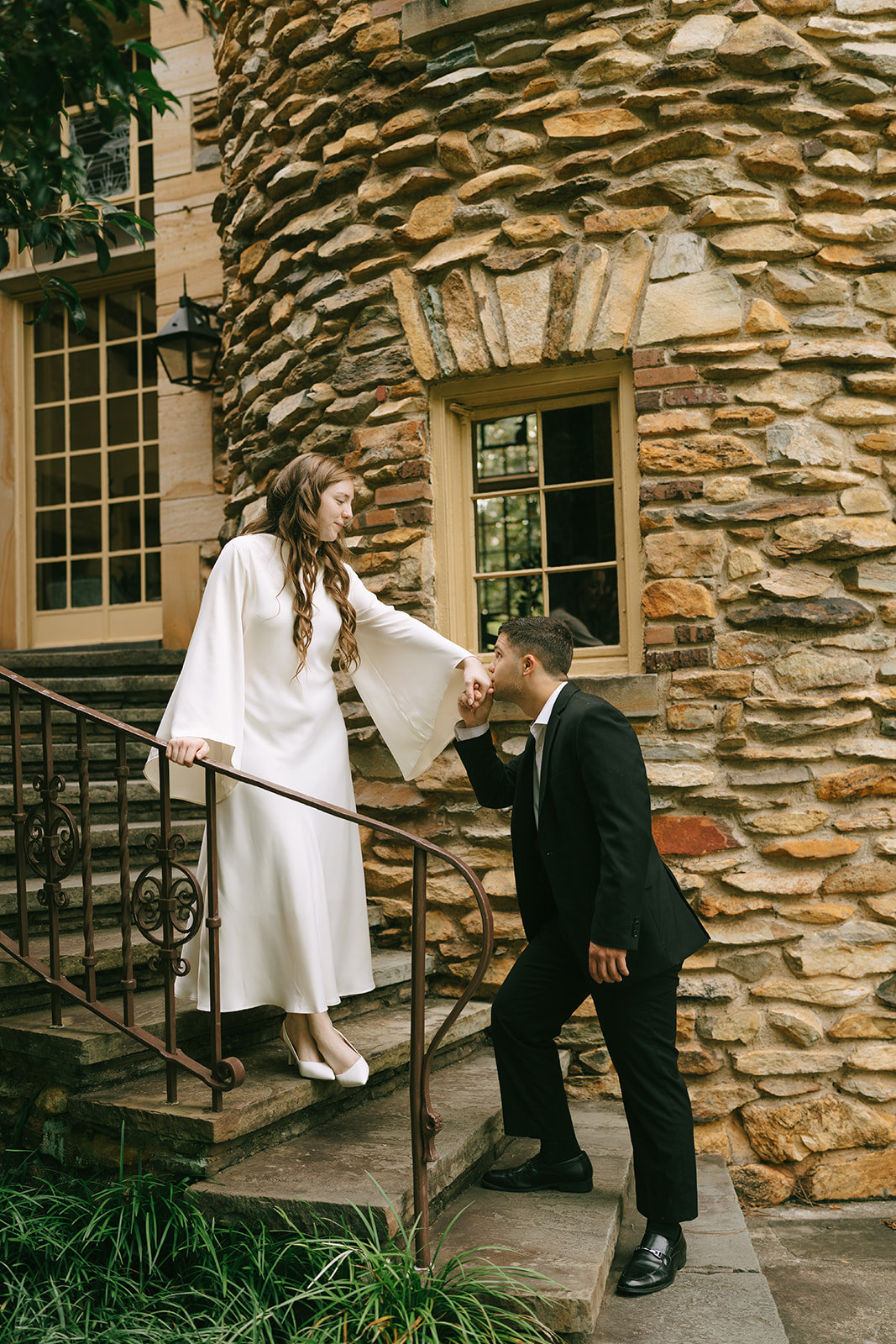 A woman in a white dress and a man in a black suit hold hands while walking on a cobblestone path in front of a stone building. | What to Wear for Engagement Photos