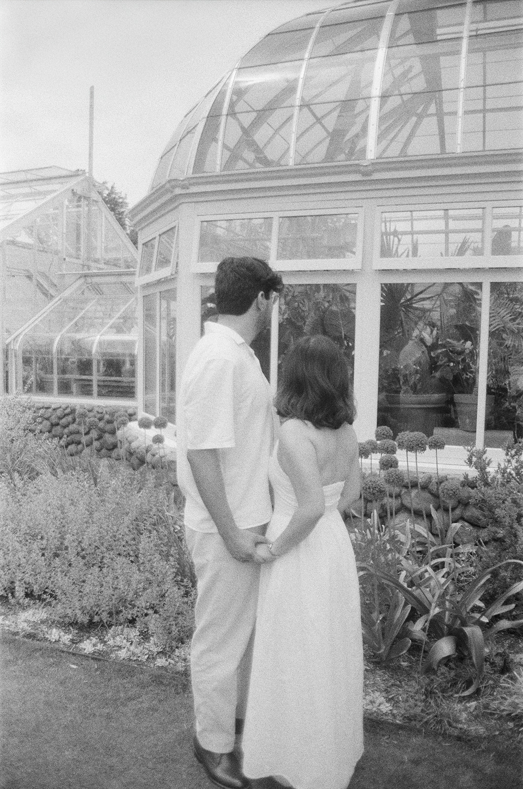 A couple stands hand in hand, facing a greenhouse surrounded by plants and flowers. | What to Wear for Engagement Photos