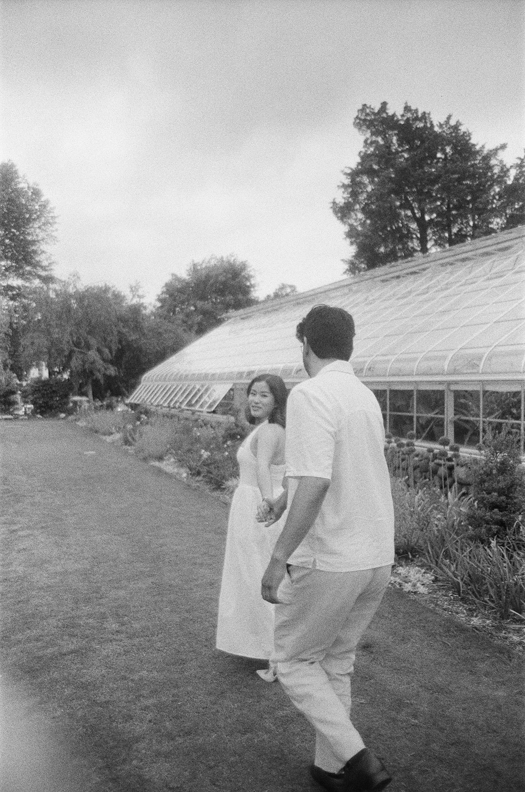 A couple stands hand in hand, facing a greenhouse surrounded by plants and flowers. | What to Wear for Engagement Photos