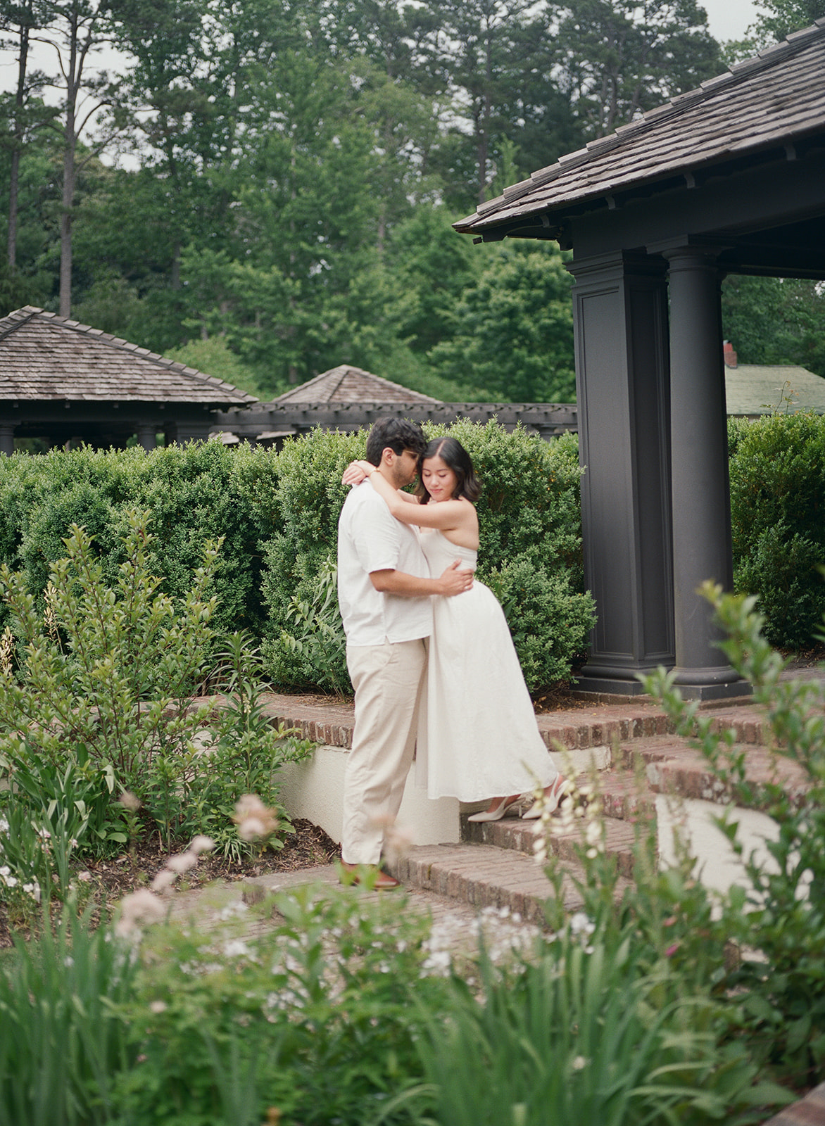 A couple stands hand in hand, facing a greenhouse surrounded by plants and flowers. | What to Wear for Engagement Photos