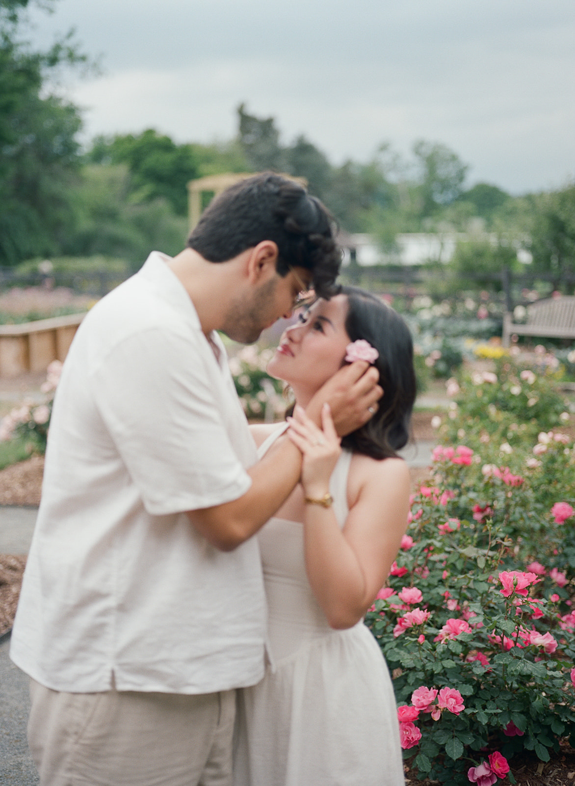 A couple dressed in white stands close together outdoors, looking into each other's eyes with greenery and flowers in the background. |What to Wear for Engagement Photos