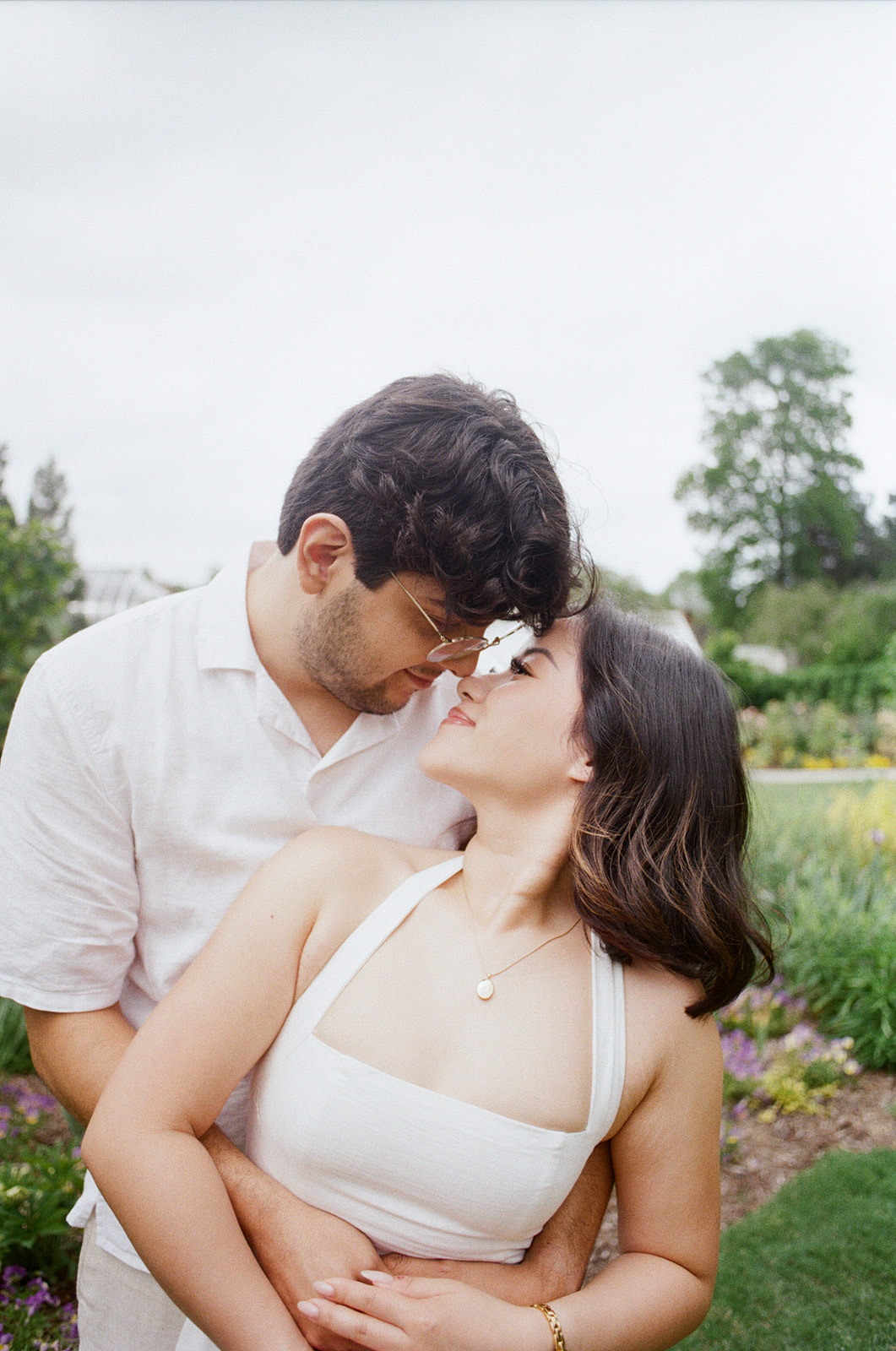A couple dressed in white stands close together outdoors, looking into each other's eyes with greenery and flowers in the background. |What to Wear for Engagement Photos