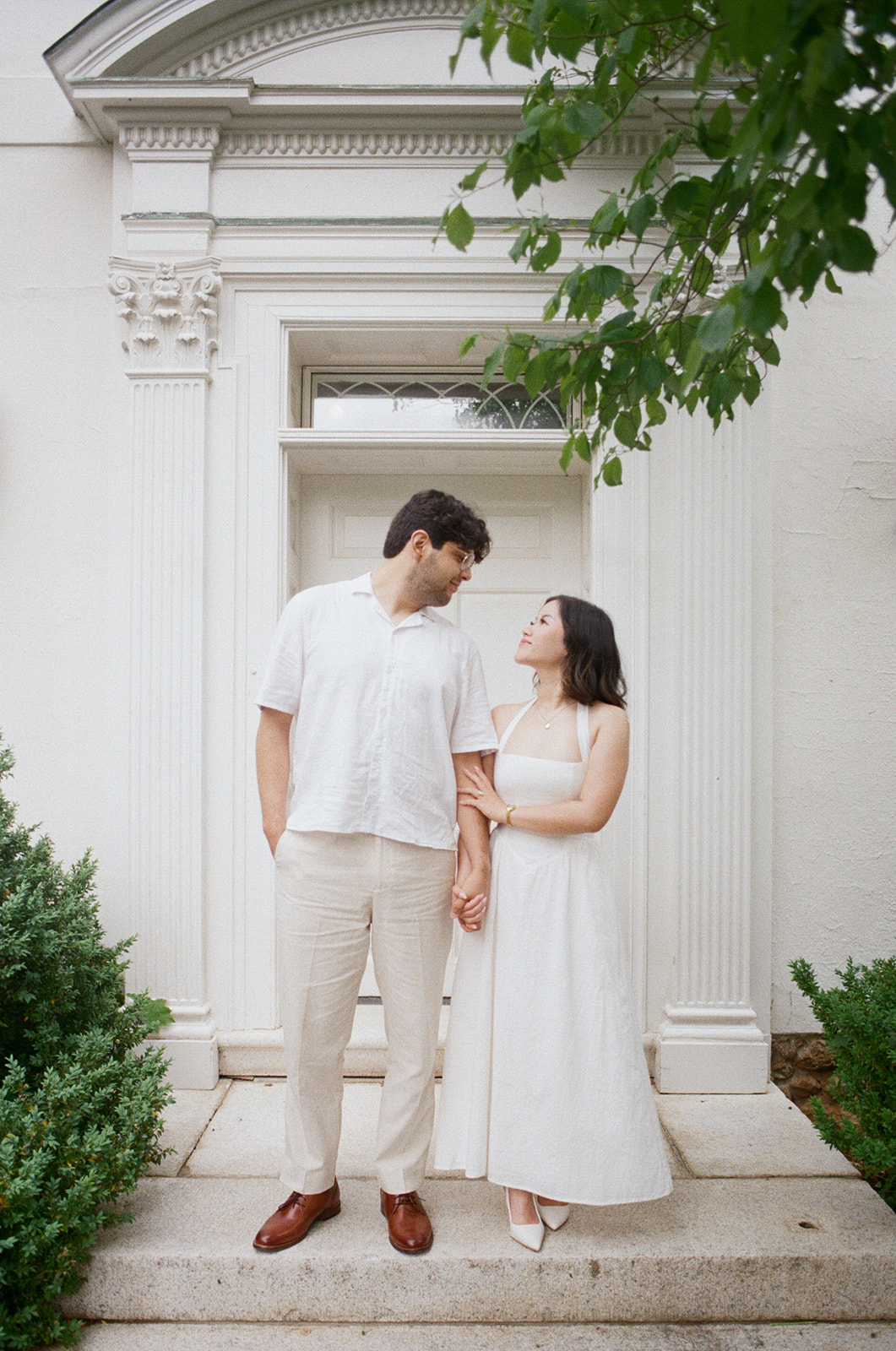A couple stands hand in hand in front of a white building with columns, both dressed in light-colored, semi-formal clothing.