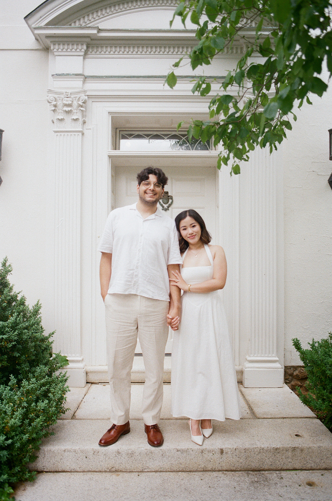 A couple stands hand in hand in front of a white building with columns, both dressed in light-colored, semi-formal clothing.