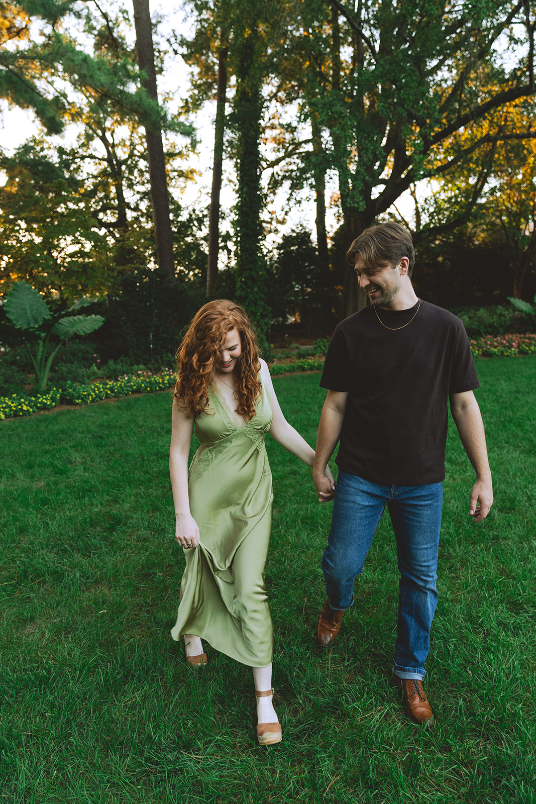 A man kisses a smiling woman on the forehead while holding her hand in a garden with trees and flowers in the background.