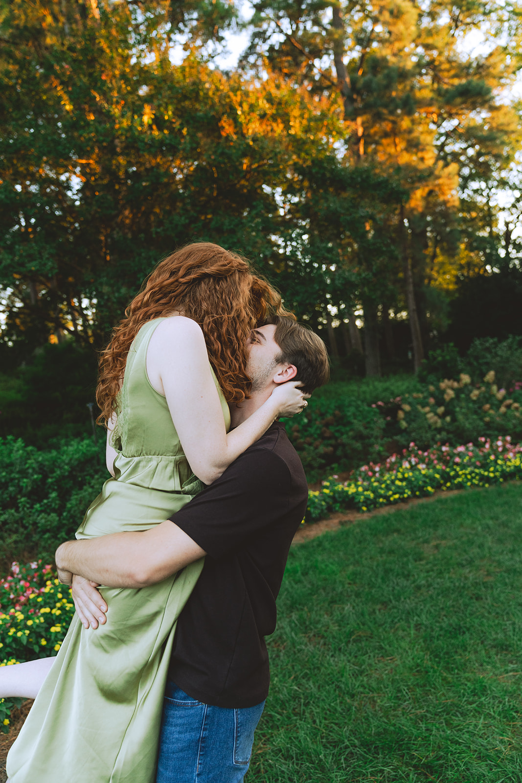 A man kisses a smiling woman on the forehead while holding her hand in a garden with trees and flowers in the background.