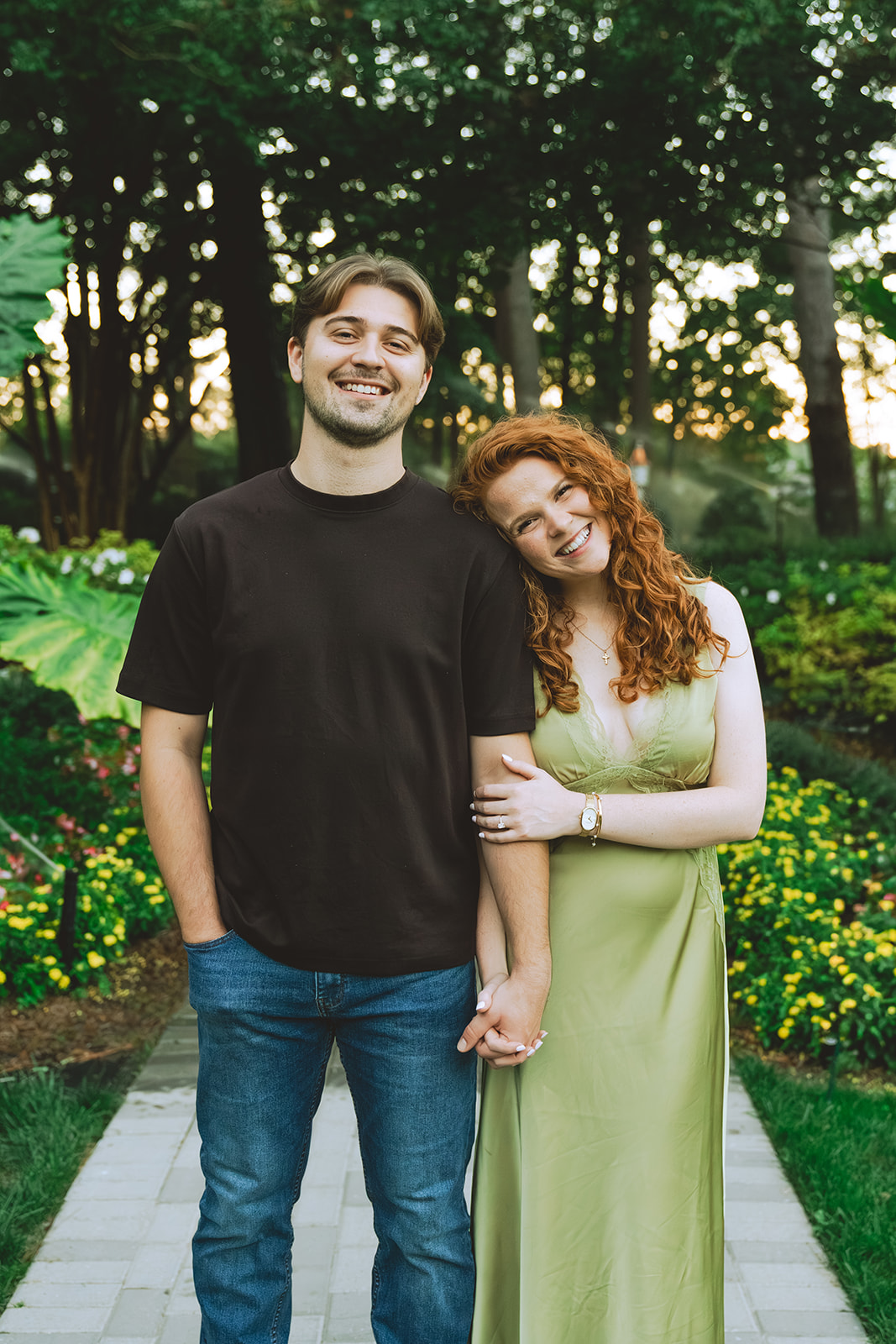 A man kisses a smiling woman on the forehead while holding her hand in a garden with trees and flowers in the background.