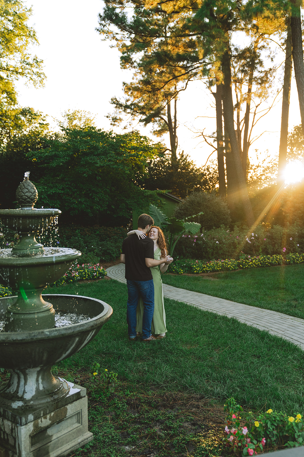 A man kisses a smiling woman on the forehead while holding her hand in a garden with trees and flowers in the background.