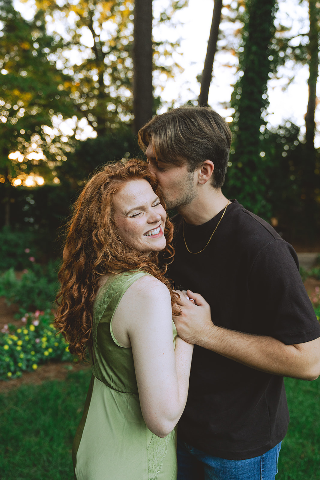 A man kisses a smiling woman on the forehead while holding her hand in a garden with trees and flowers in the background.