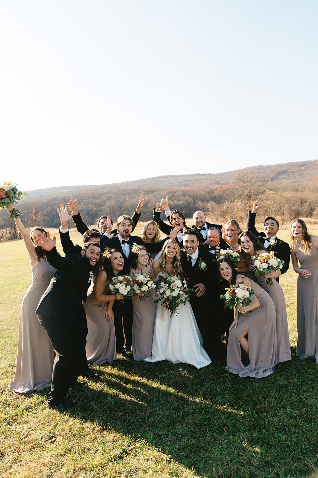 A wedding party poses outdoors in formal attire, with bridesmaids in taupe dresses and groomsmen in black suits, smiling and raising their arms on a grassy field.