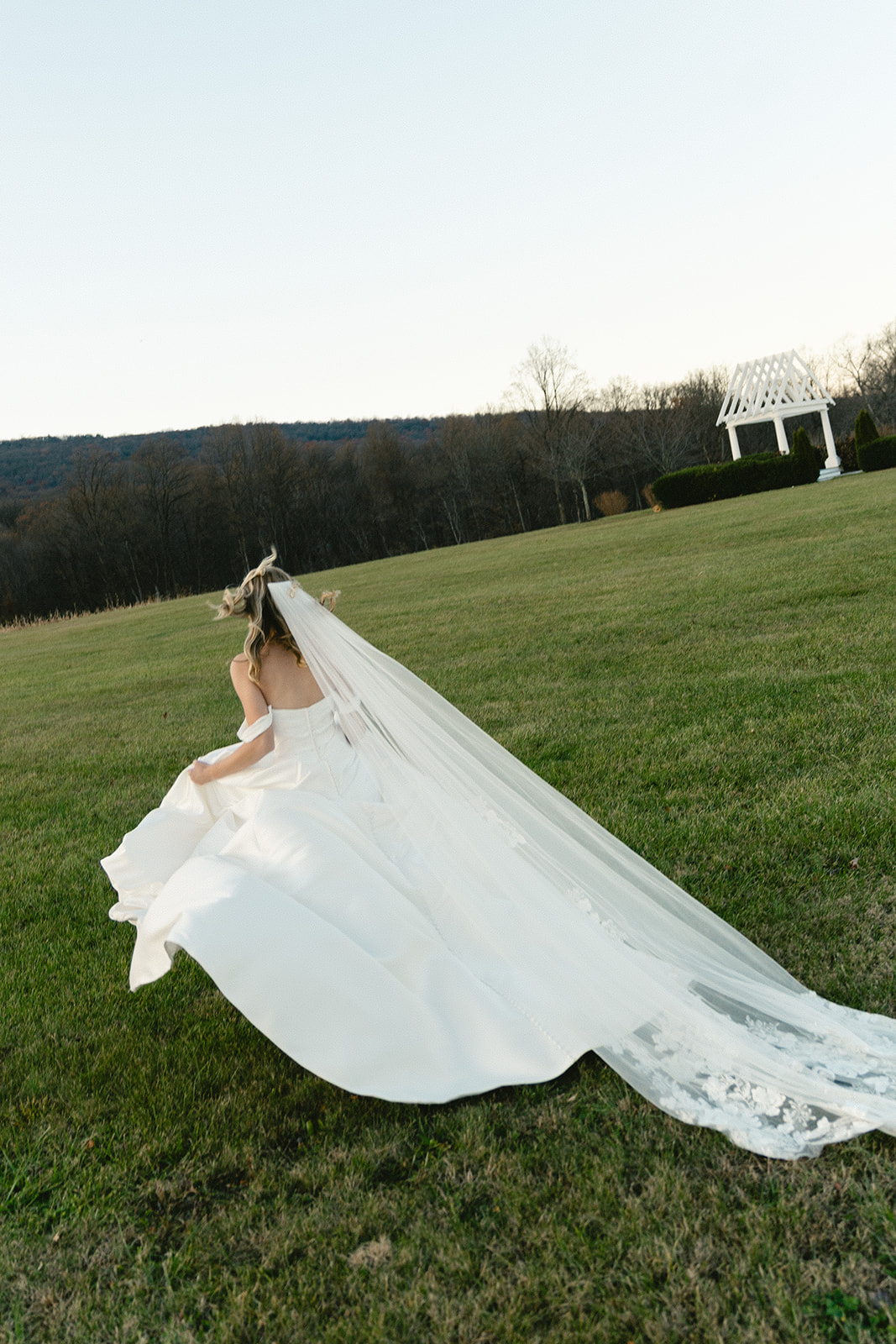 A bride in a white wedding dress and long veil walks across a grassy field toward a white gazebo under a clear sky.