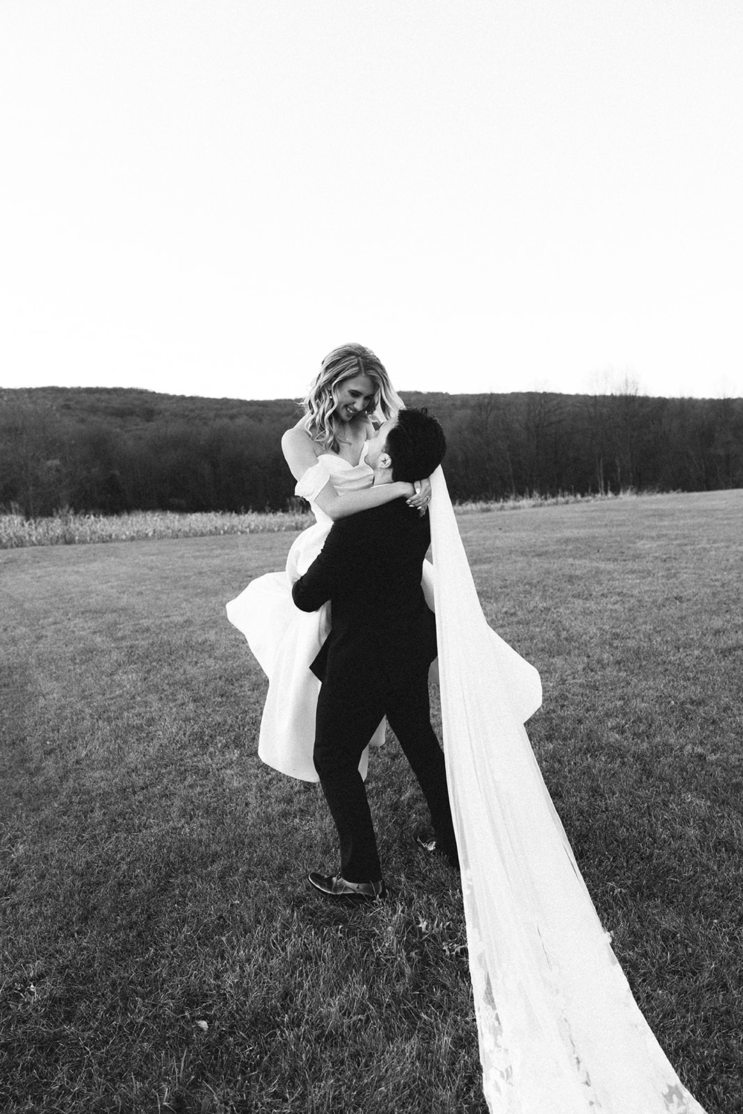 A person in a suit carries a person in a wedding dress with a long train across a grassy field. The scene is outdoors with hills and trees in the background.