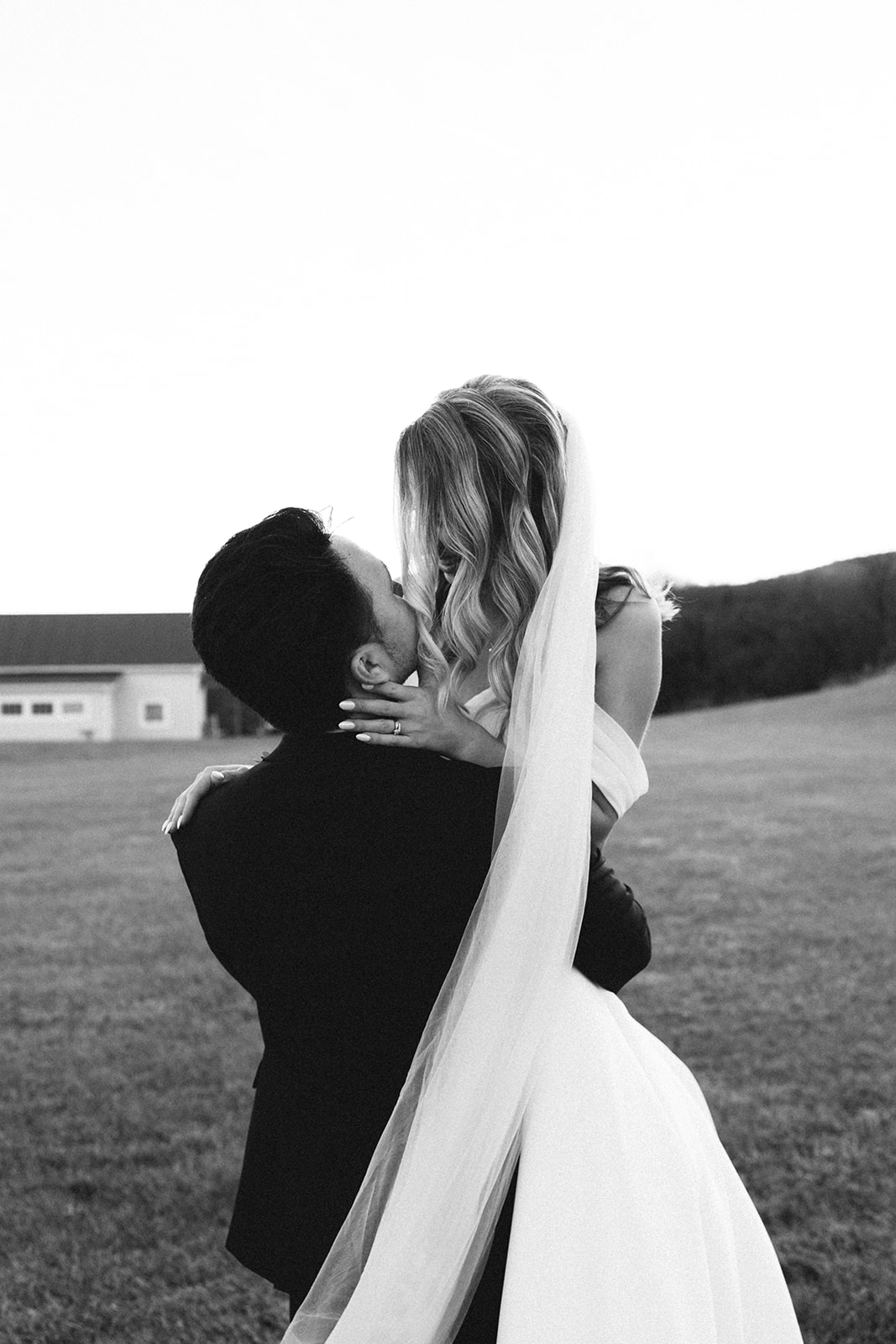 A bride and groom in formal attire stand outdoors, embracing. The bride wears an off-shoulder dress; the groom has a boutonniere and suit. The photo is in black and white.