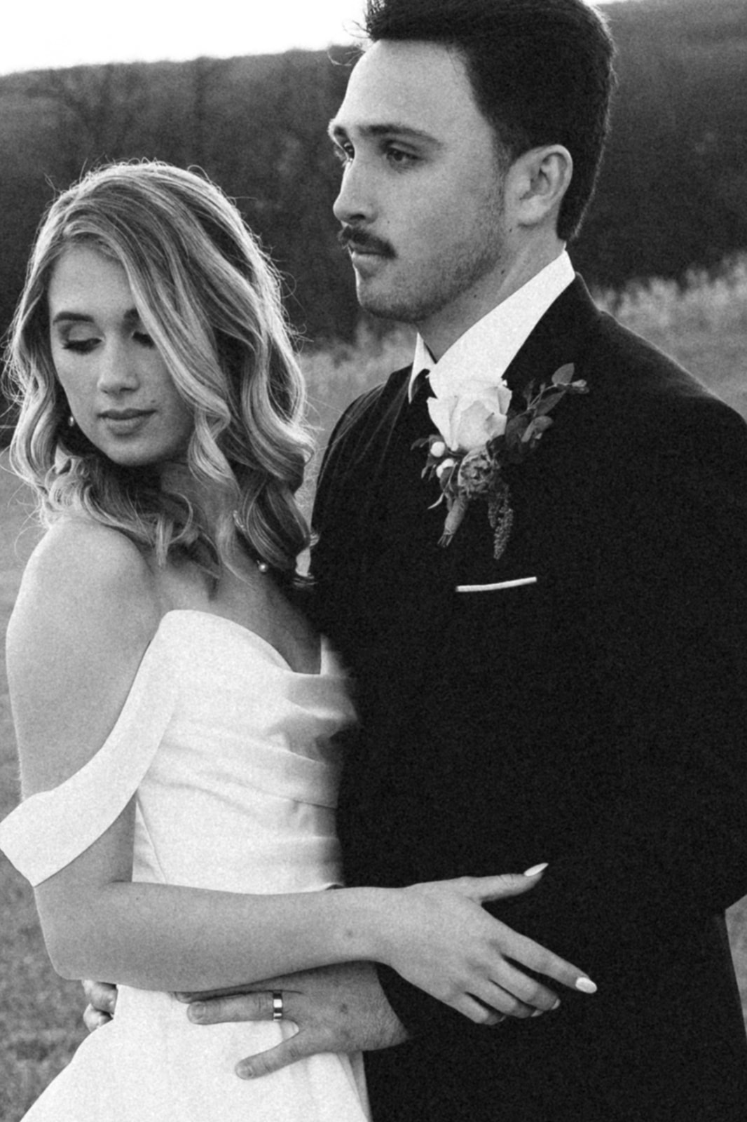 A bride and groom in formal attire stand outdoors, embracing. The bride wears an off-shoulder dress; the groom has a boutonniere and suit. The photo is in black and white.