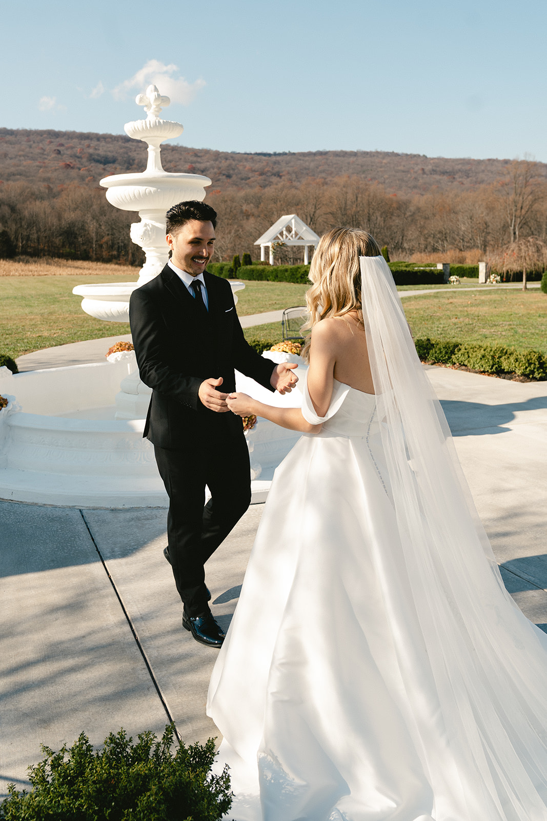 A bride in a white gown approaches a groom standing ahead near a gazebo and fountain in an outdoor setting for their first look at their wedding