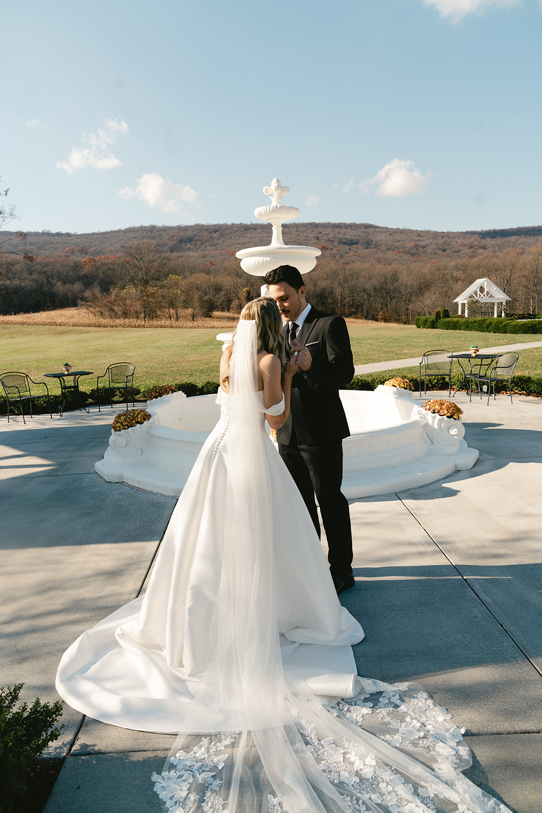 A bride in a white gown approaches a groom standing ahead near a gazebo and fountain in an outdoor setting for their first look at their wedding