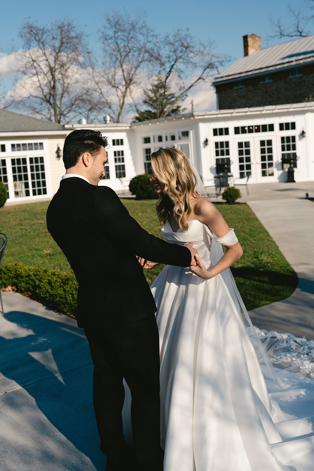 A bride in a white gown approaches a groom standing ahead near a gazebo and fountain in an outdoor setting for their first look at their wedding