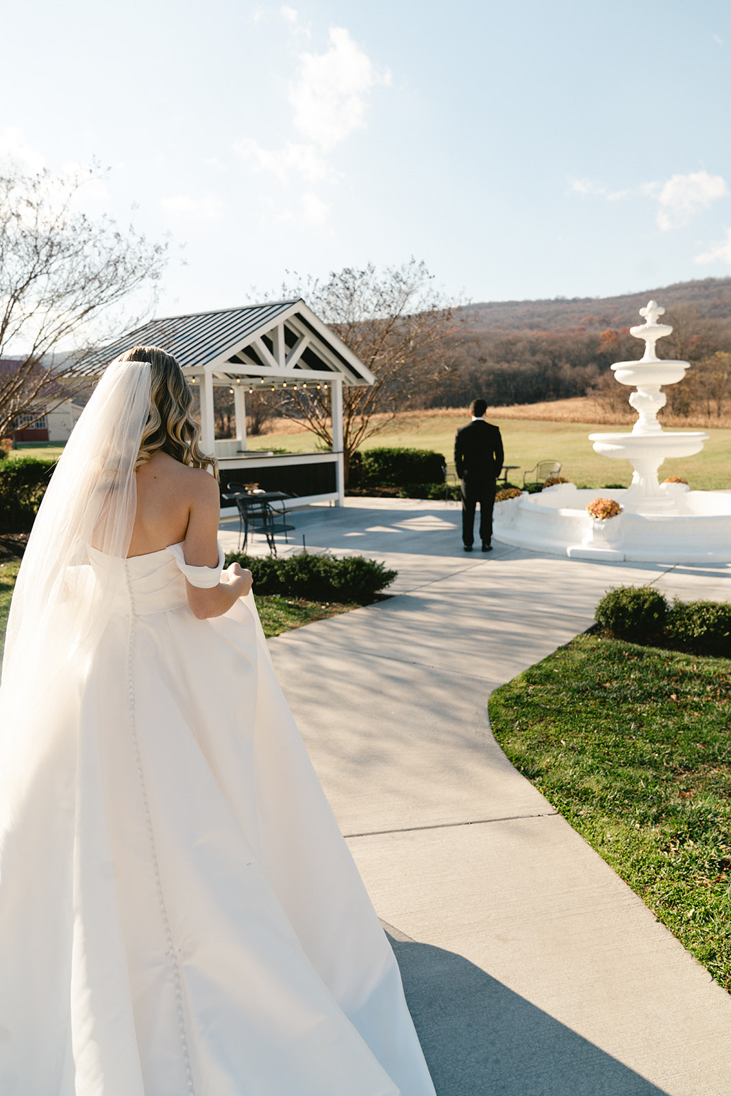 A bride in a white gown approaches a groom standing ahead near a gazebo and fountain in an outdoor setting for their first look at their wedding