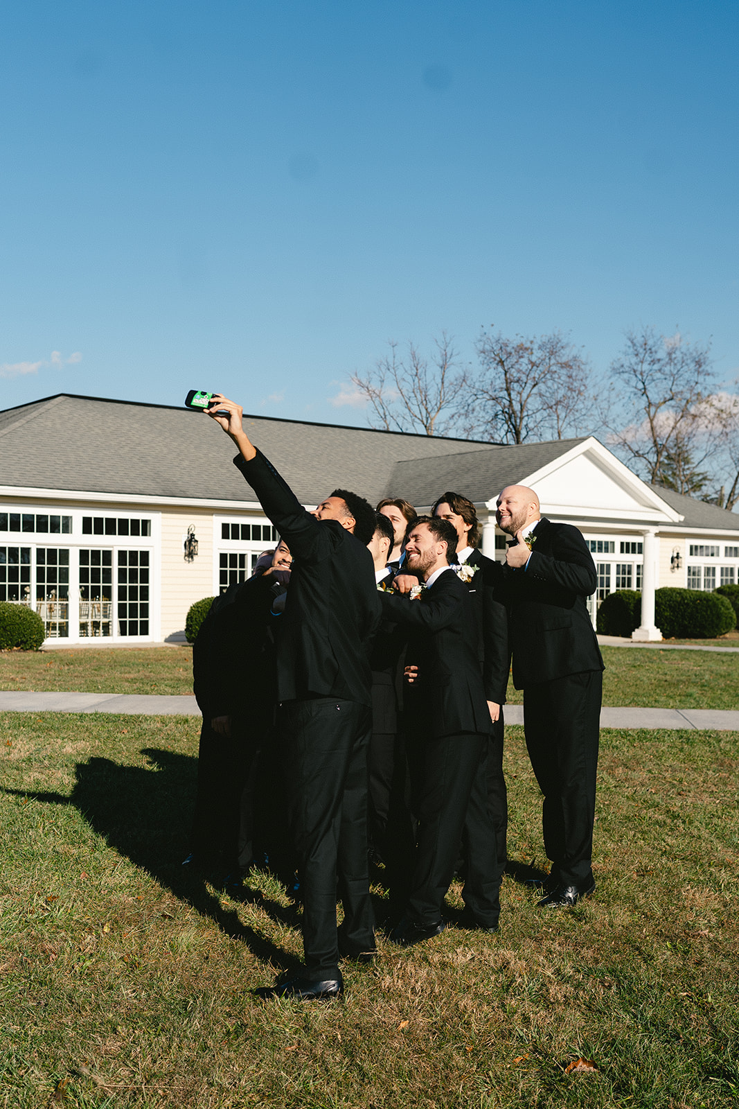 A group of people in formal black attire stand on grass outside a building, gathered closely together while taking a selfie.