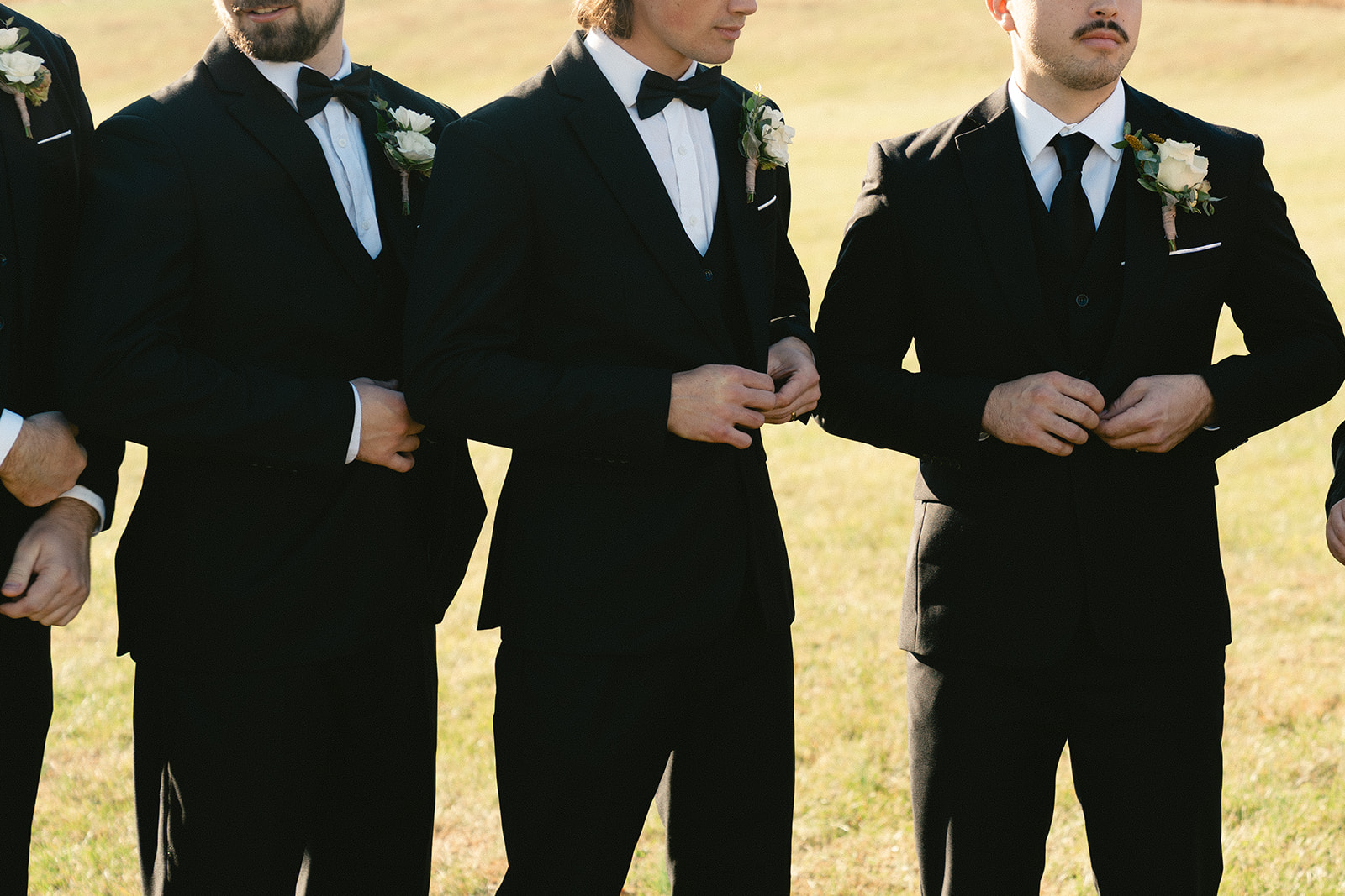 Three men in black suits with bow ties and boutonnieres stand outdoors, adjusting their jackets. Only their upper bodies are visible.