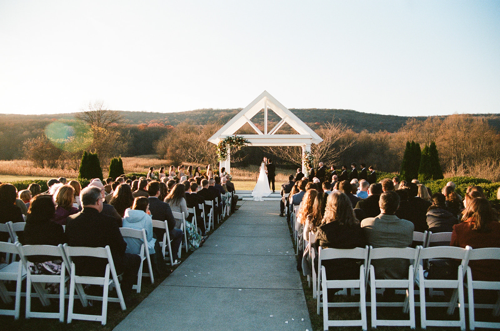 Outdoor wedding ceremony with guests seated on either side of an aisle, facing a couple standing under a white wooden arch.