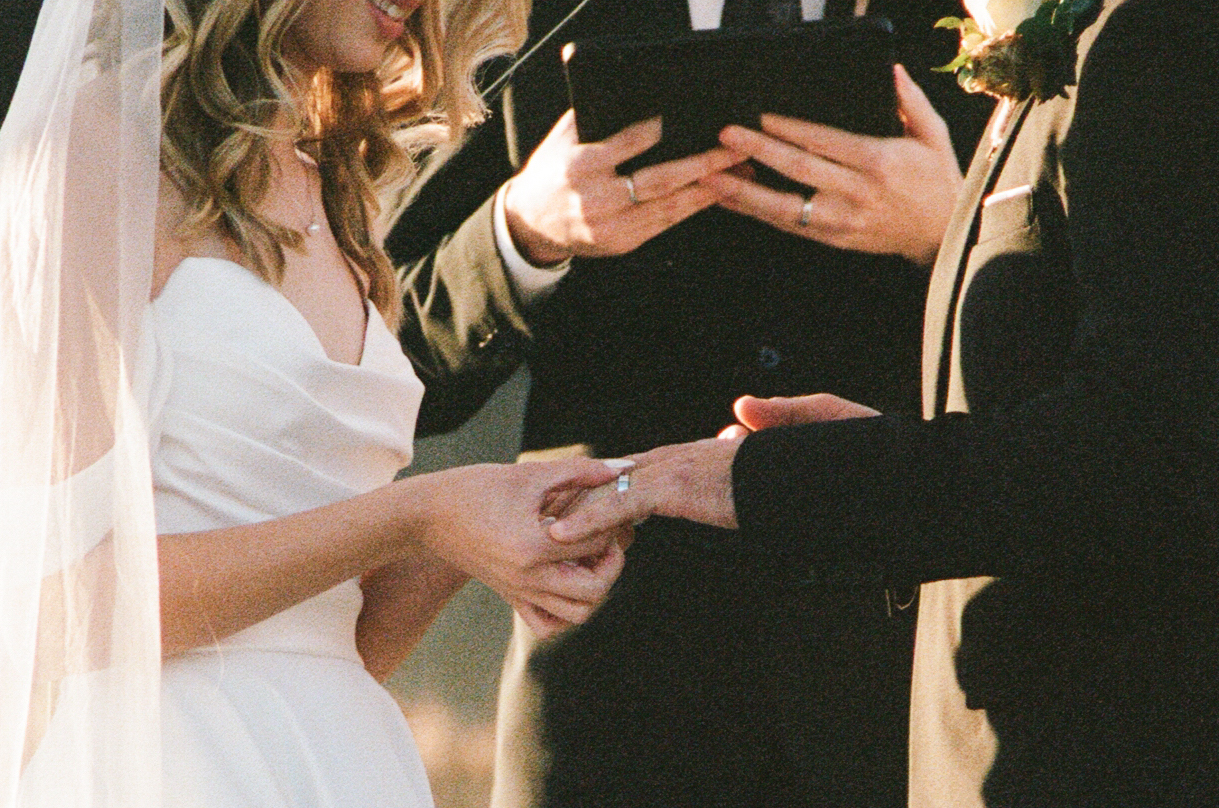 A bride and groom exchange rings during their wedding ceremony, with an officiant holding a book in the background.