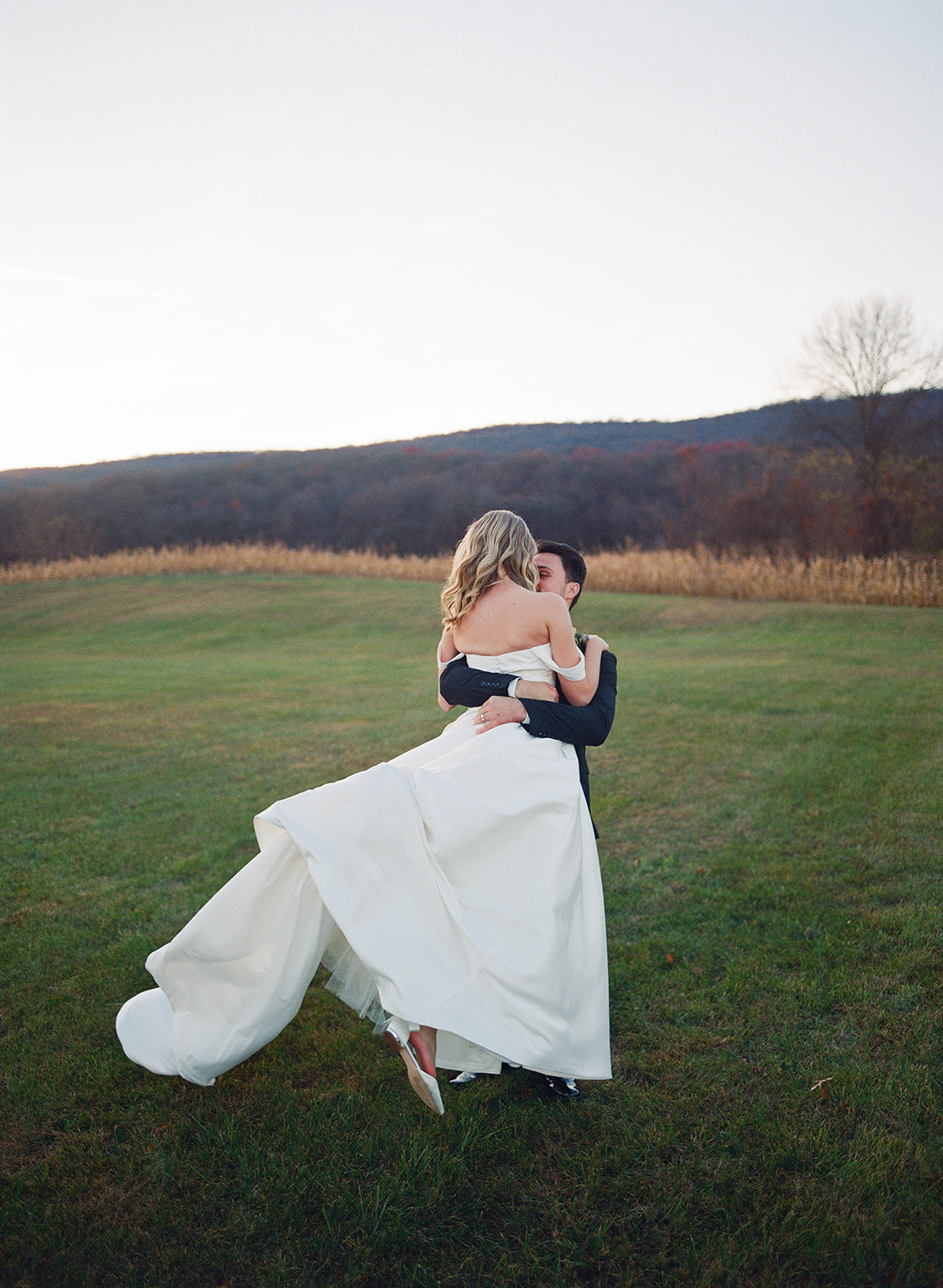A person in a suit carries a person in a wedding dress with a long train across a grassy field. The scene is outdoors with hills and trees in the background.