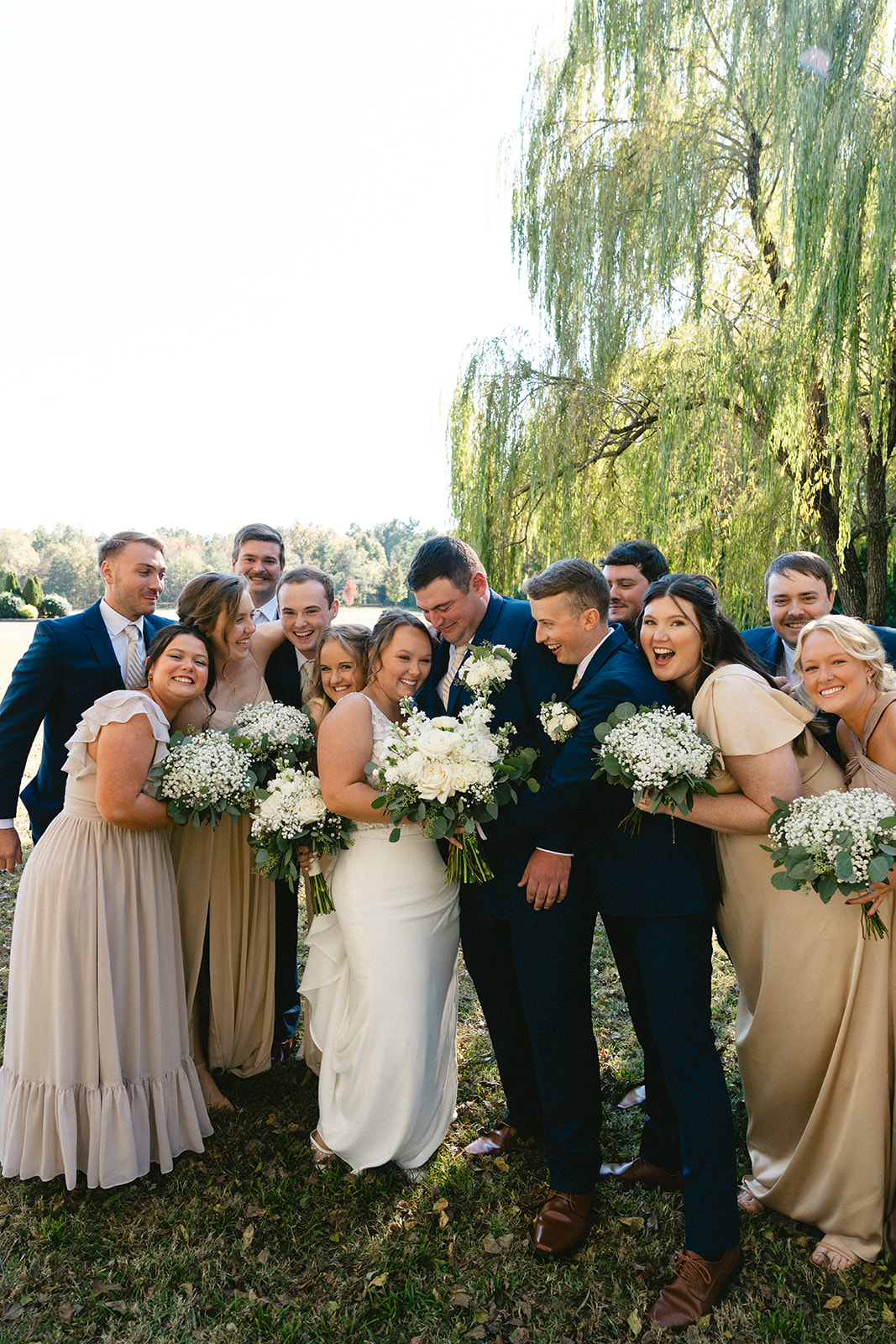 Five women in formal dresses, including a bride in white holding a bouquet, stand together outdoors smiling, with greenery and trees in the background. | How to Choose a Wedding Photographer