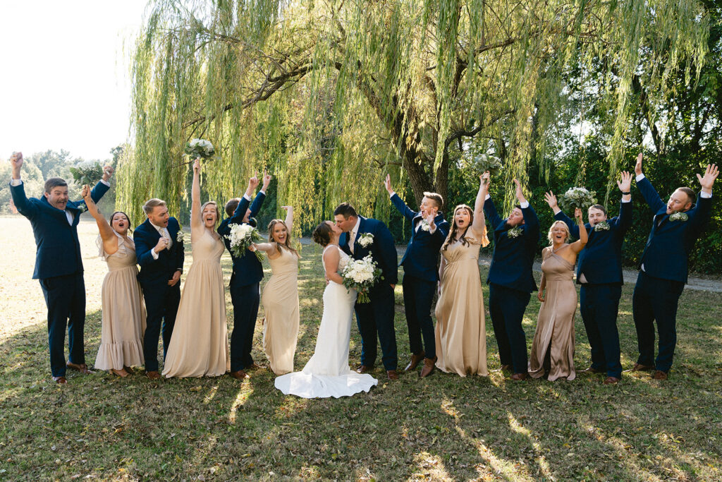 A wedding party poses outdoors under a large willow tree, with the bride and groom in the center and attendants raising their arms and smiling.