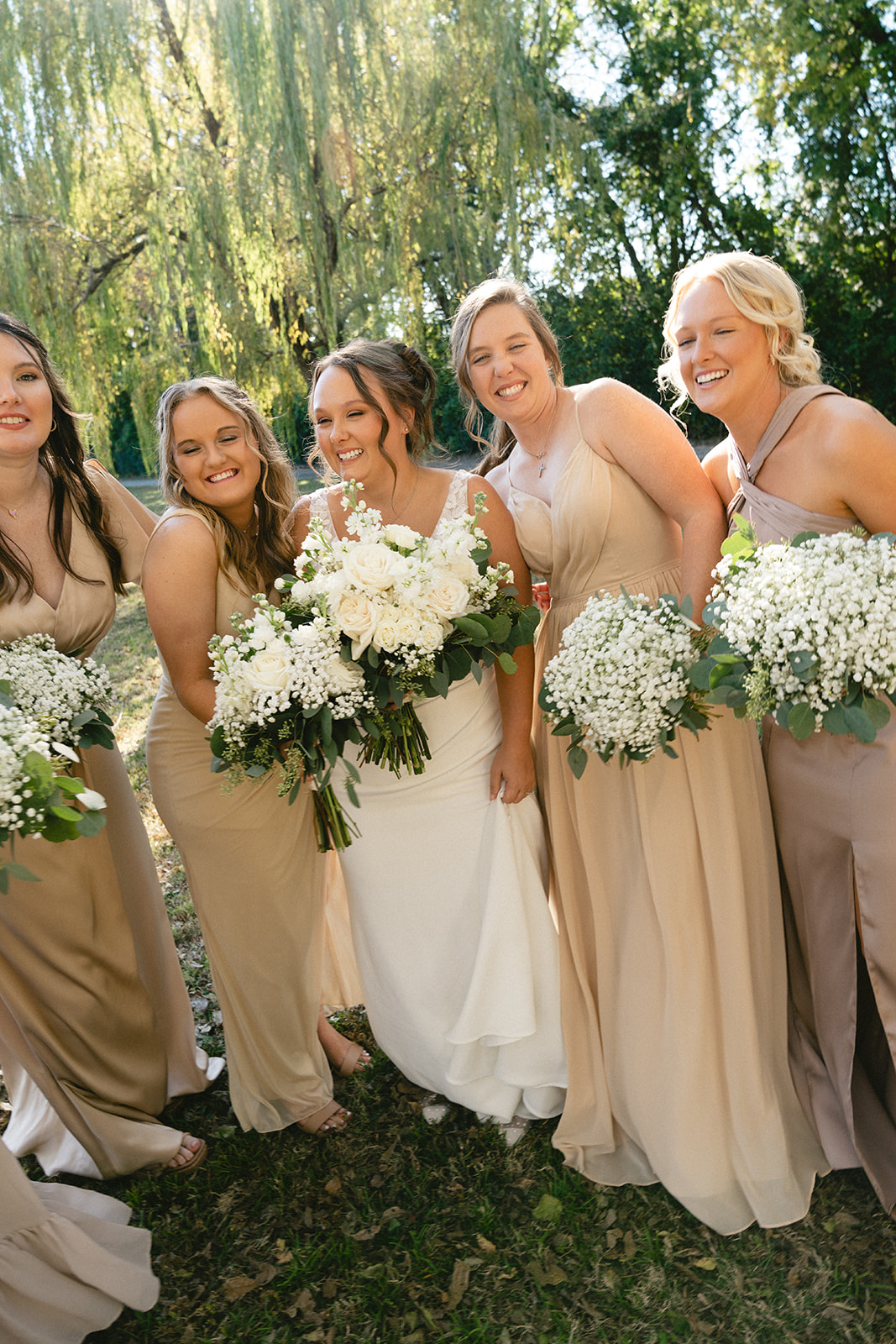 Five women in formal dresses, including a bride in white holding a bouquet, stand together outdoors smiling, with greenery and trees in the background. | How to Choose a Wedding Photographer