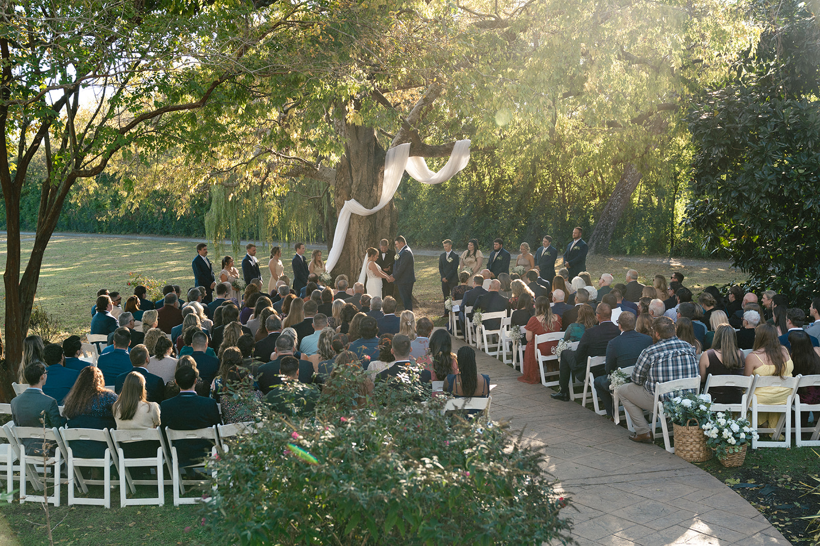 Outdoor wedding ceremony with guests seated on white chairs facing a couple and officiant under a large tree decorated with white fabric, in daylight.
