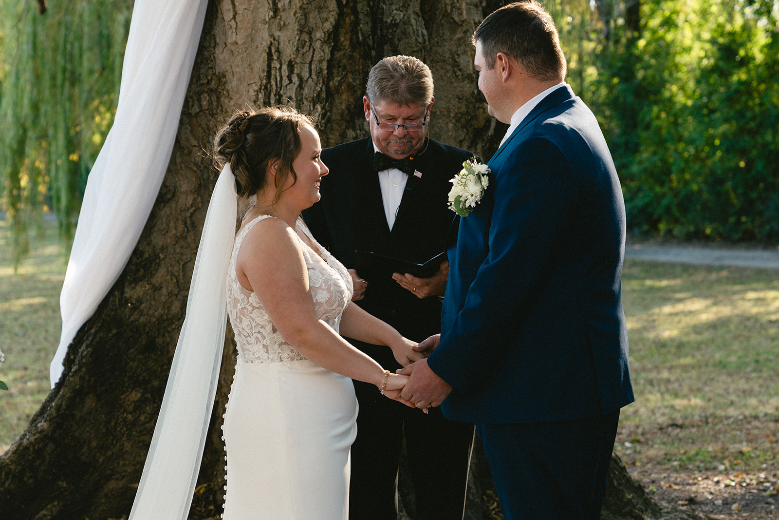 A bride and groom stand holding hands during an outdoor wedding ceremony, with an officiant reading from a book behind them.