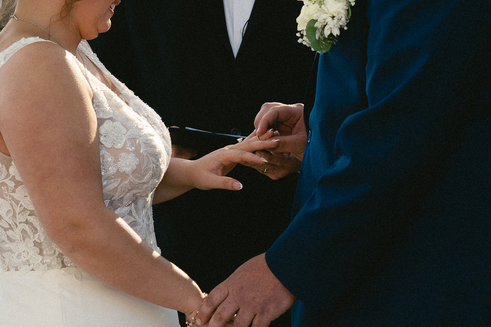 A bride and groom stand holding hands during an outdoor wedding ceremony, with an officiant reading from a book behind them.