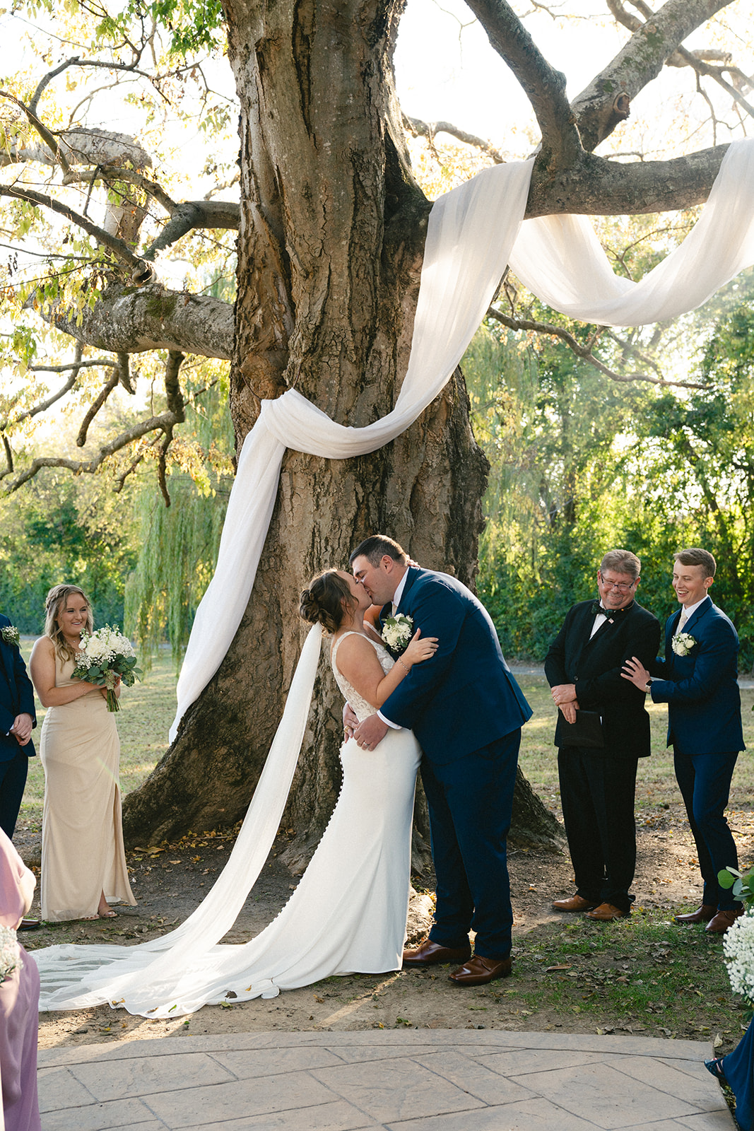A bride and groom share a kiss during their outdoor wedding ceremony, with a large tree draped in white fabric and guests standing nearby. | How to Choose a Wedding Photographer