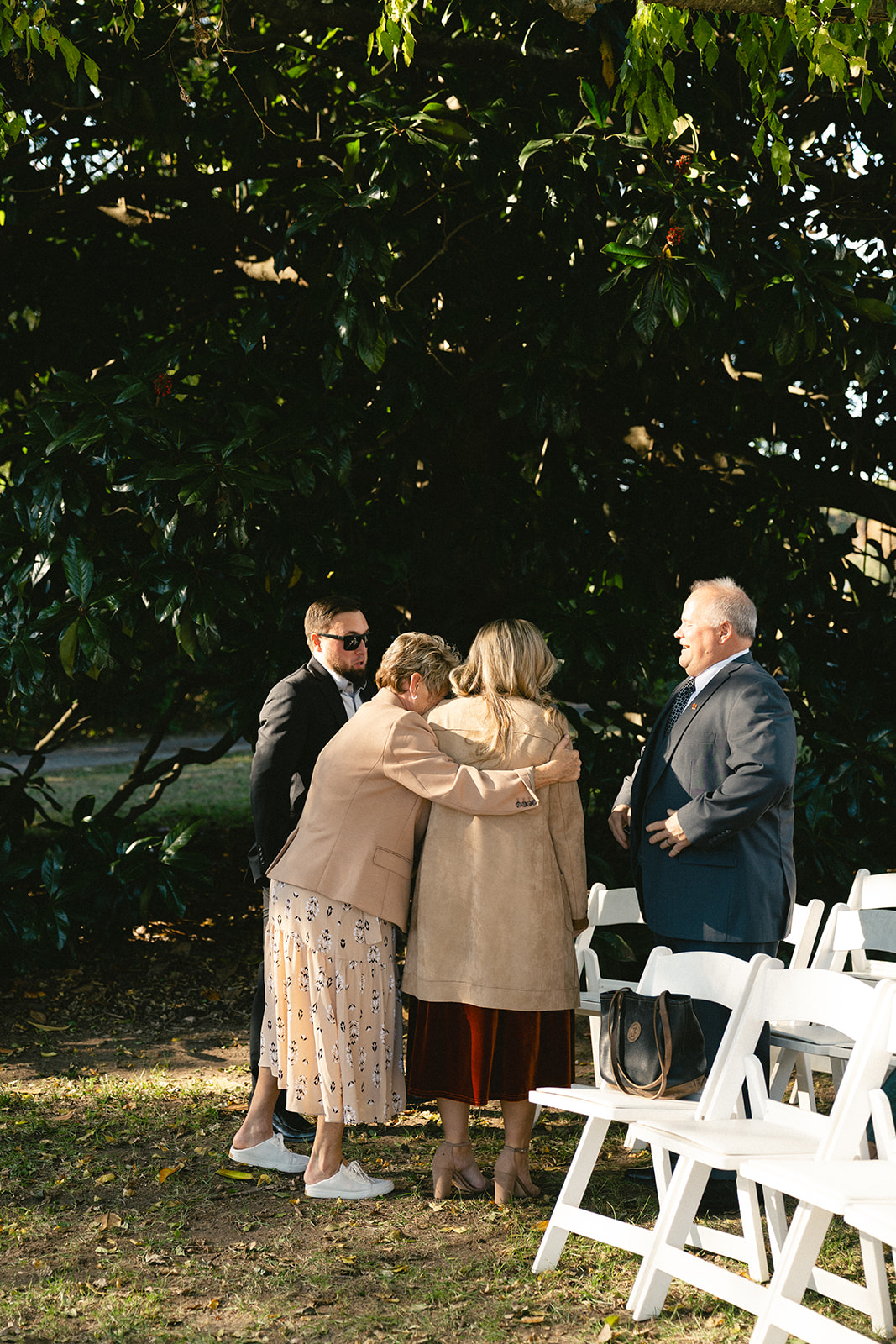 Four adults stand and converse near white folding chairs outdoors, with two women embracing and two men wearing suits under a large tree.