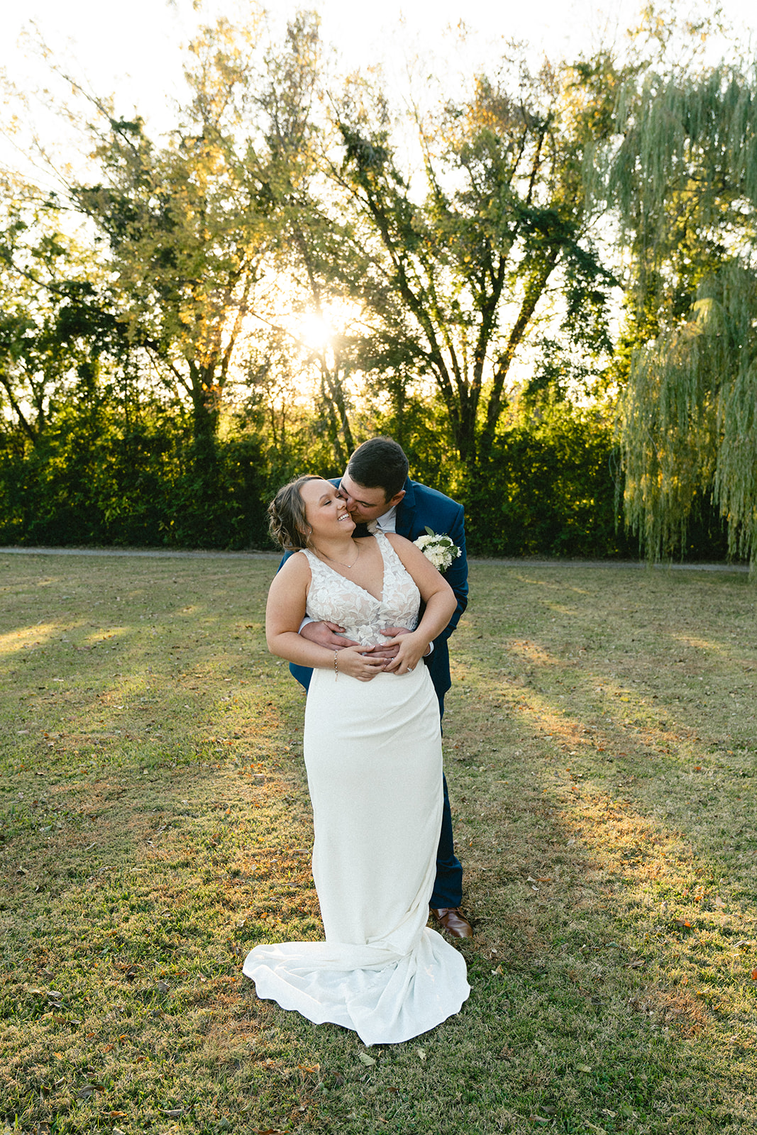 A bride and groom stand together outdoors on a grassy field, holding hands and looking at the camera, with trees in the background. | How to Choose a Wedding Photographer