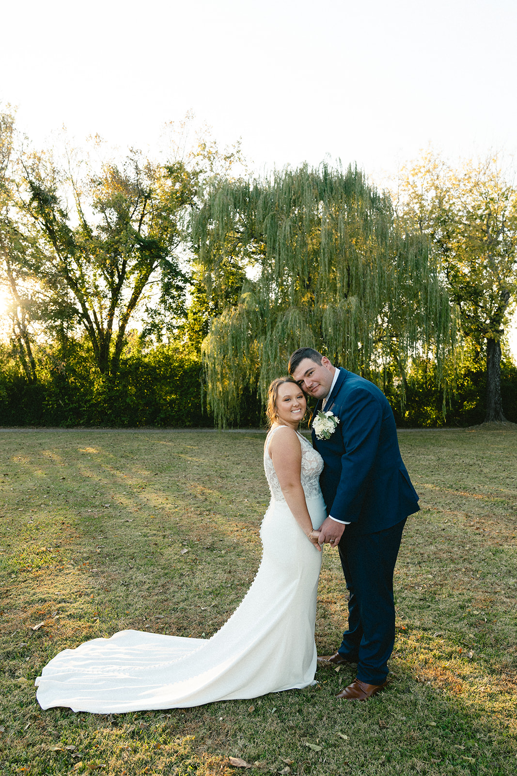 A bride and groom stand together outdoors on a grassy field, holding hands and looking at the camera, with trees in the background. | How to Choose a Wedding Photographer