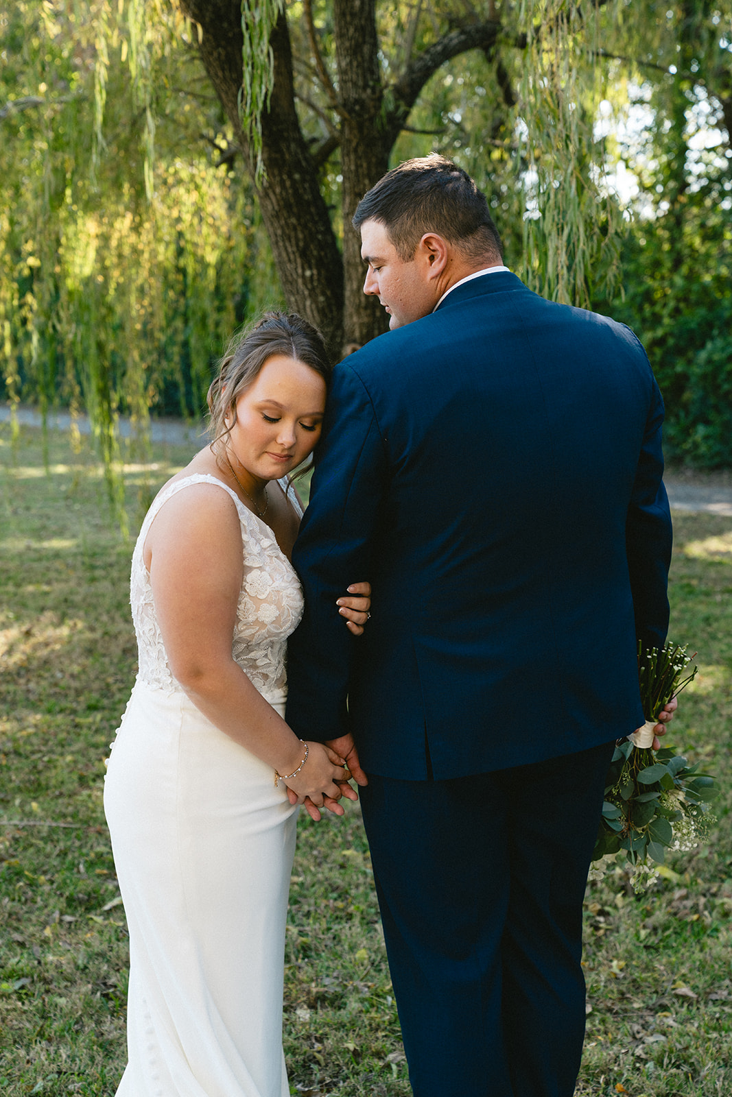 A bride and groom stand together outdoors on a grassy field, holding hands and looking at the camera, with trees in the background. | How to Choose a Wedding Photographer