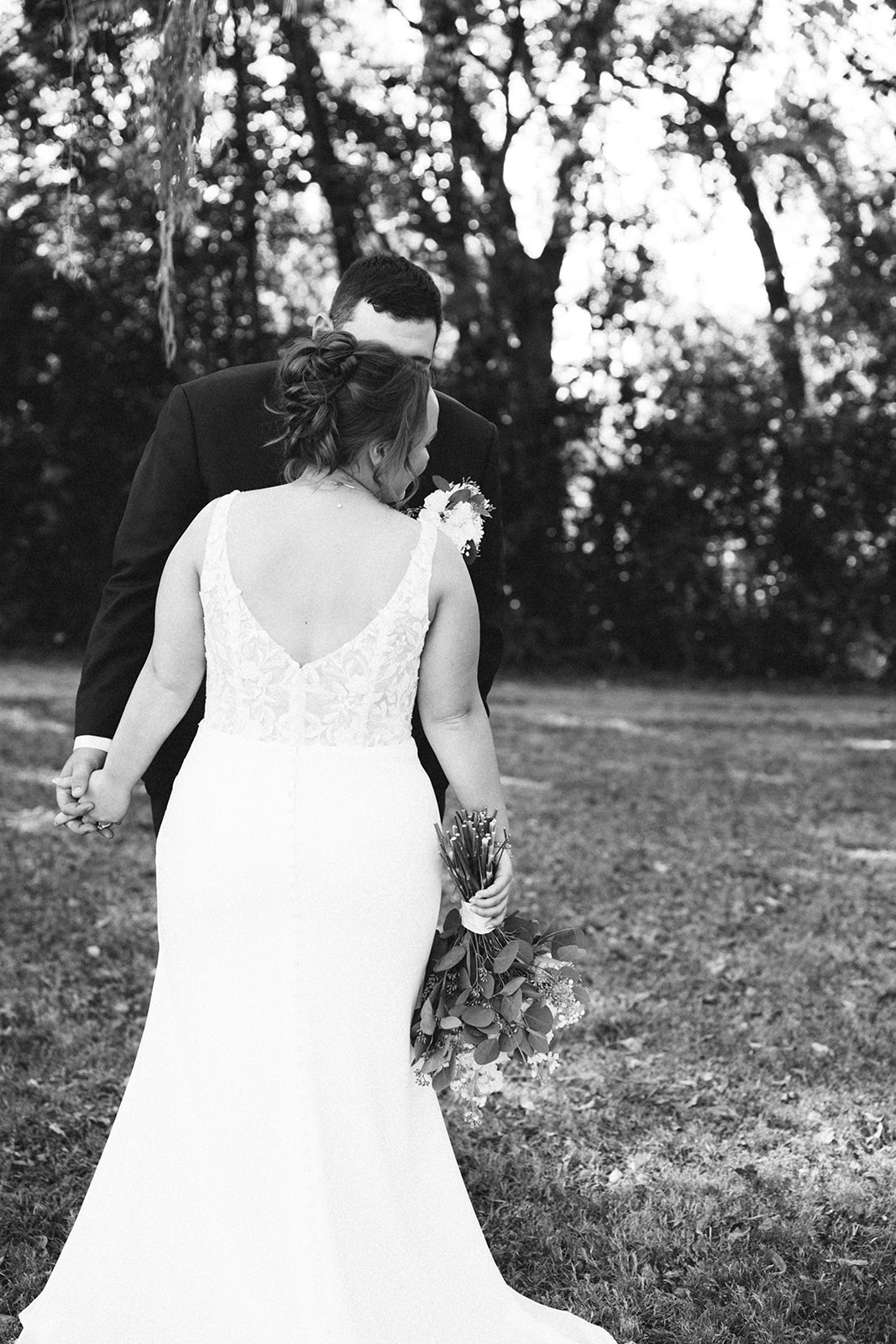 A bride and groom stand together outdoors on a grassy field, holding hands and looking at the camera, with trees in the background. | How to Choose a Wedding Photographer